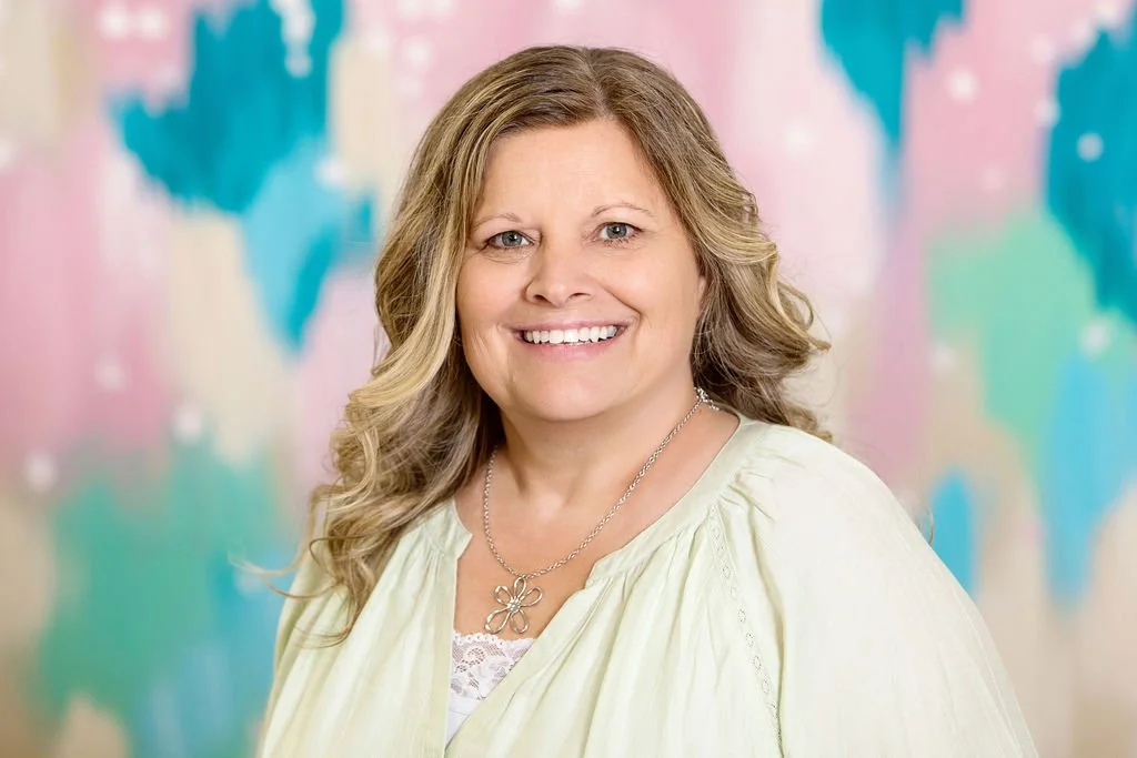 A woman with long, curly blonde hair smiling for a portrait in front of a colorful abstract background.