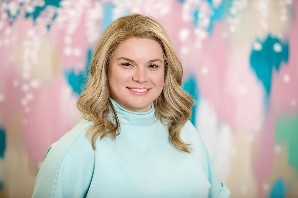 A woman with blonde hair in a white blazer and light jeans standing in front of a colorful mural of pink and white cherry blossoms on a blue background.