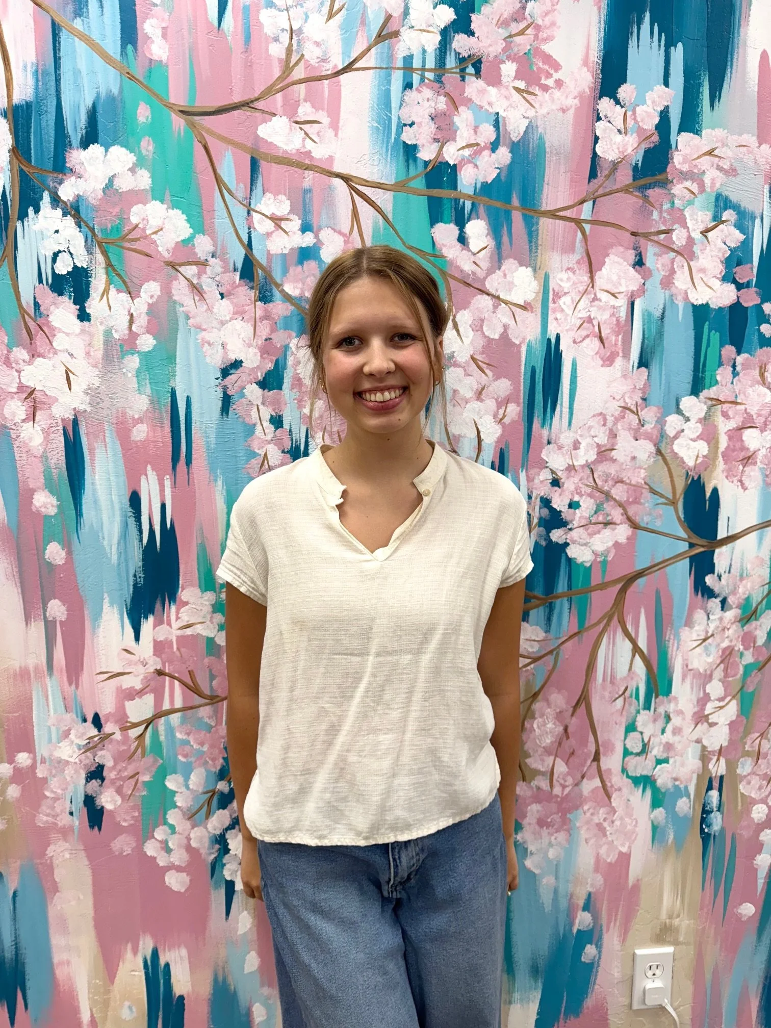 A young woman with light brown hair tied back, wearing a cream-colored short-sleeve blouse and blue jeans, standing in front of a colorful mural of pink cherry blossoms on abstract blue and pink backgrounds.