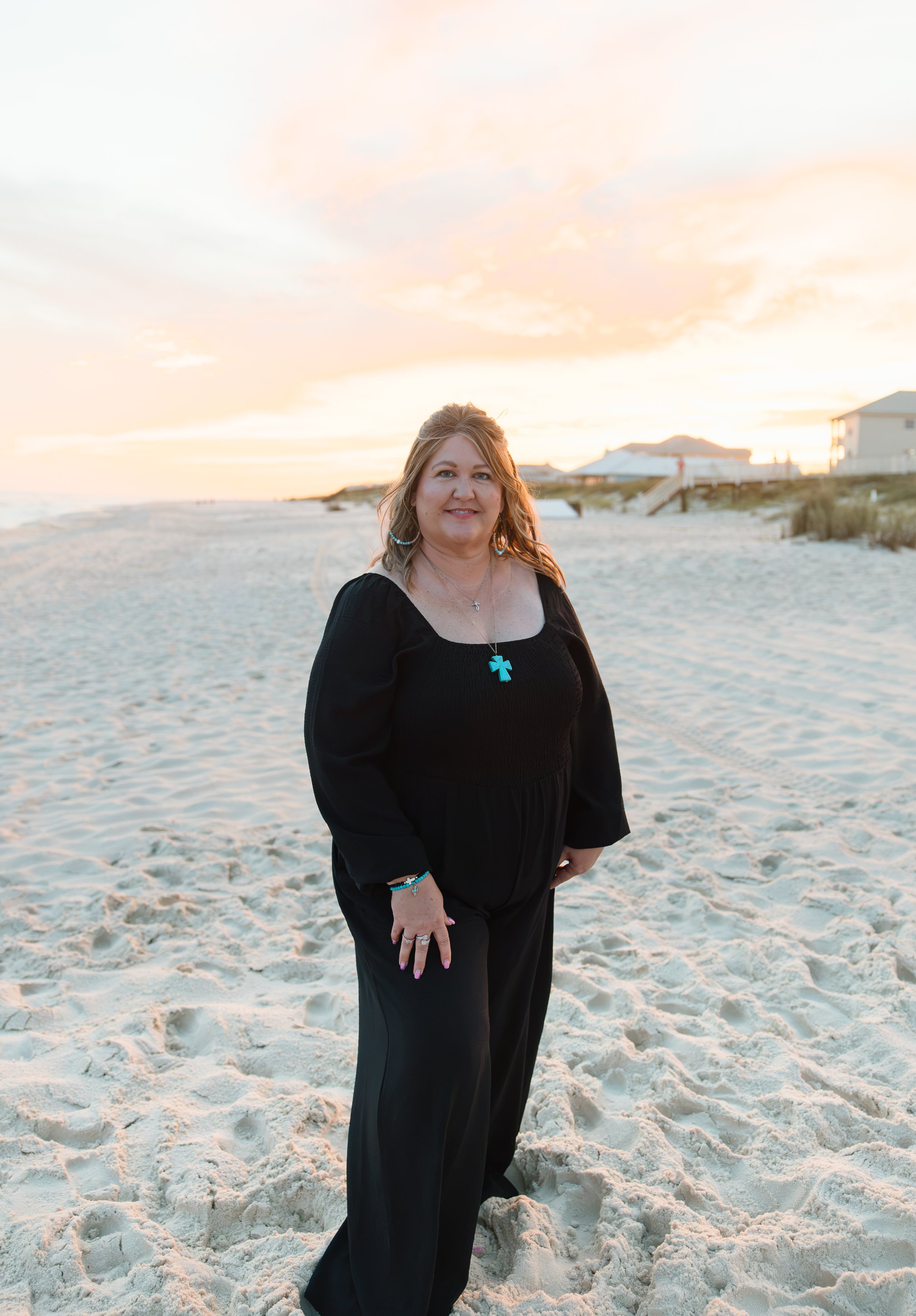 A woman stands on a sandy beach during sunset, wearing a black dress and smiling at the camera.