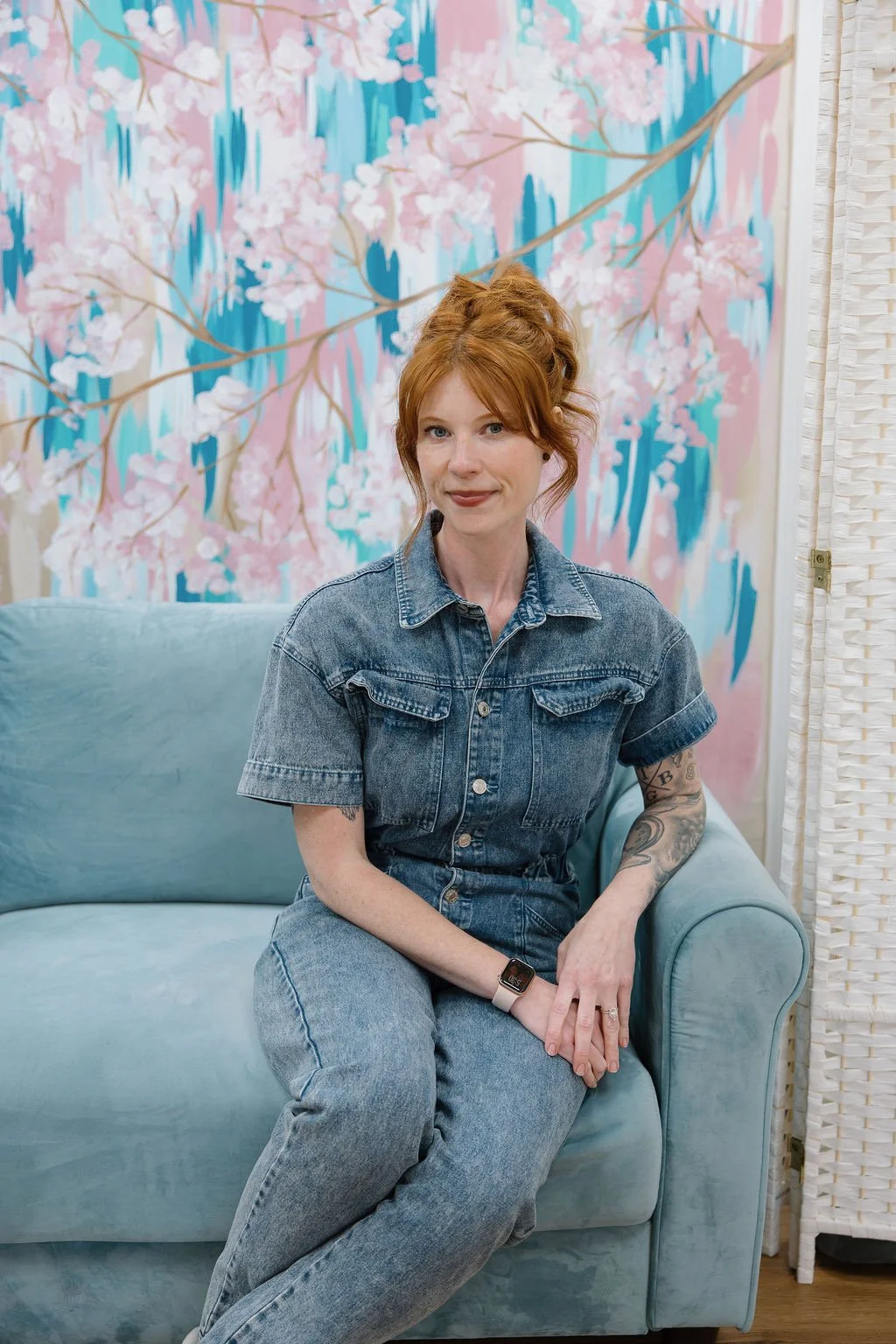 A woman with red hair in a white shirt and blue jeans standing in front of a colorful mural of pink cherry blossoms with blue and pink abstract background.