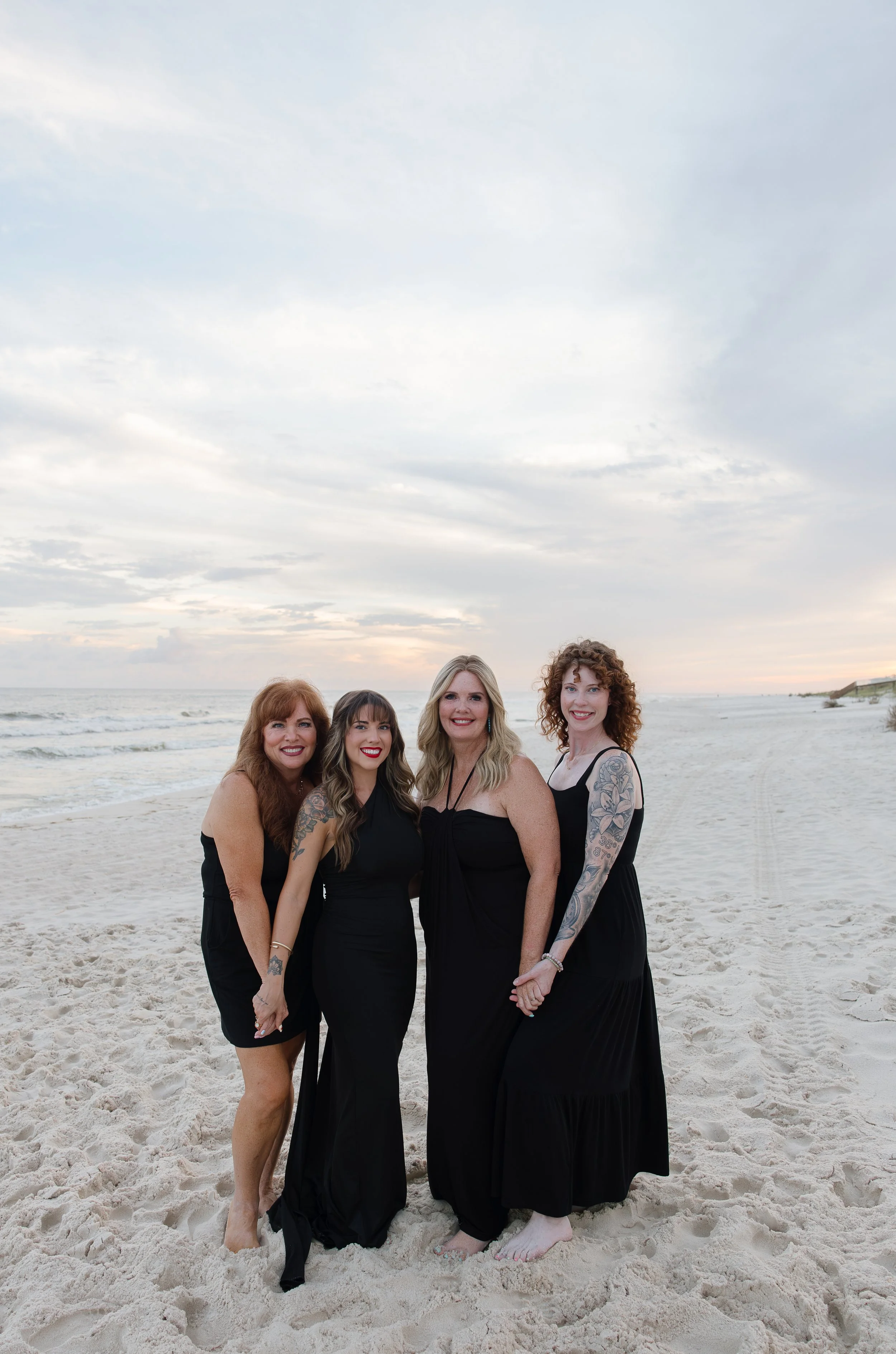 Group of five women standing on a sandy beach with ocean waves and cloudy sky in the background. They are dressed in black dresses and smiling at the camera.