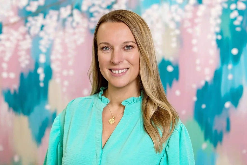 A woman with long blonde hair wearing a light turquoise blouse and a gold necklace, standing in front of a colorful abstract background with pink, blue, and white hues.