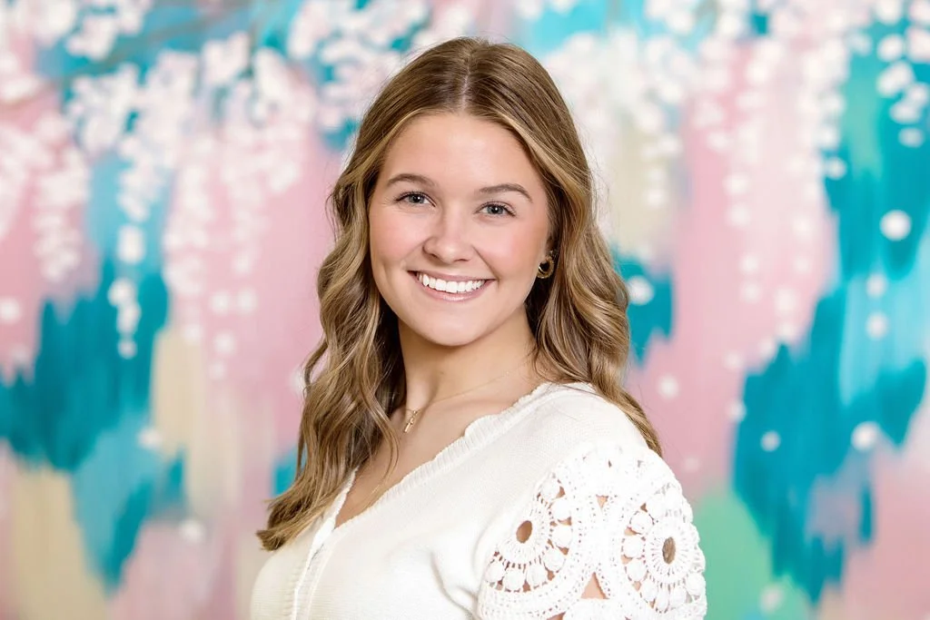 A young woman with wavy light brown hair, wearing a white shirt and blue jeans, sitting on a beige couch with a blue pillow, smiling at the camera in a plain room.