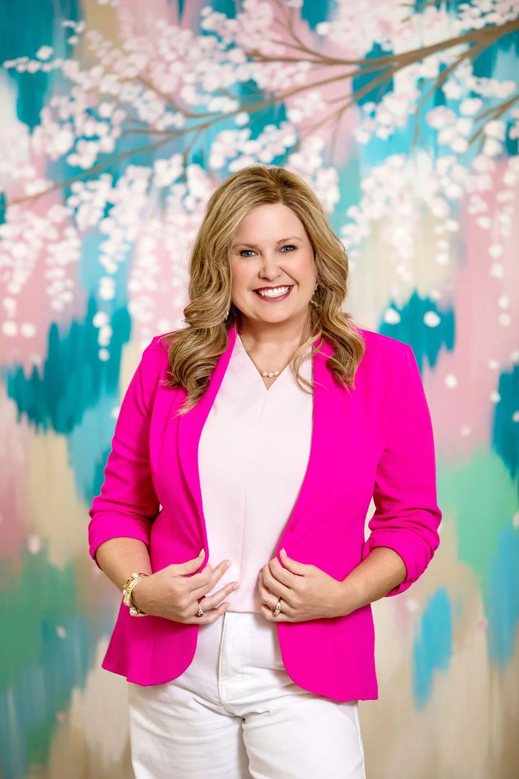 A woman with wavy blonde hair wearing a bright pink blazer, white top, and white pants, standing in front of a colorful background with pastel pink, blue, and green abstract floral design.
