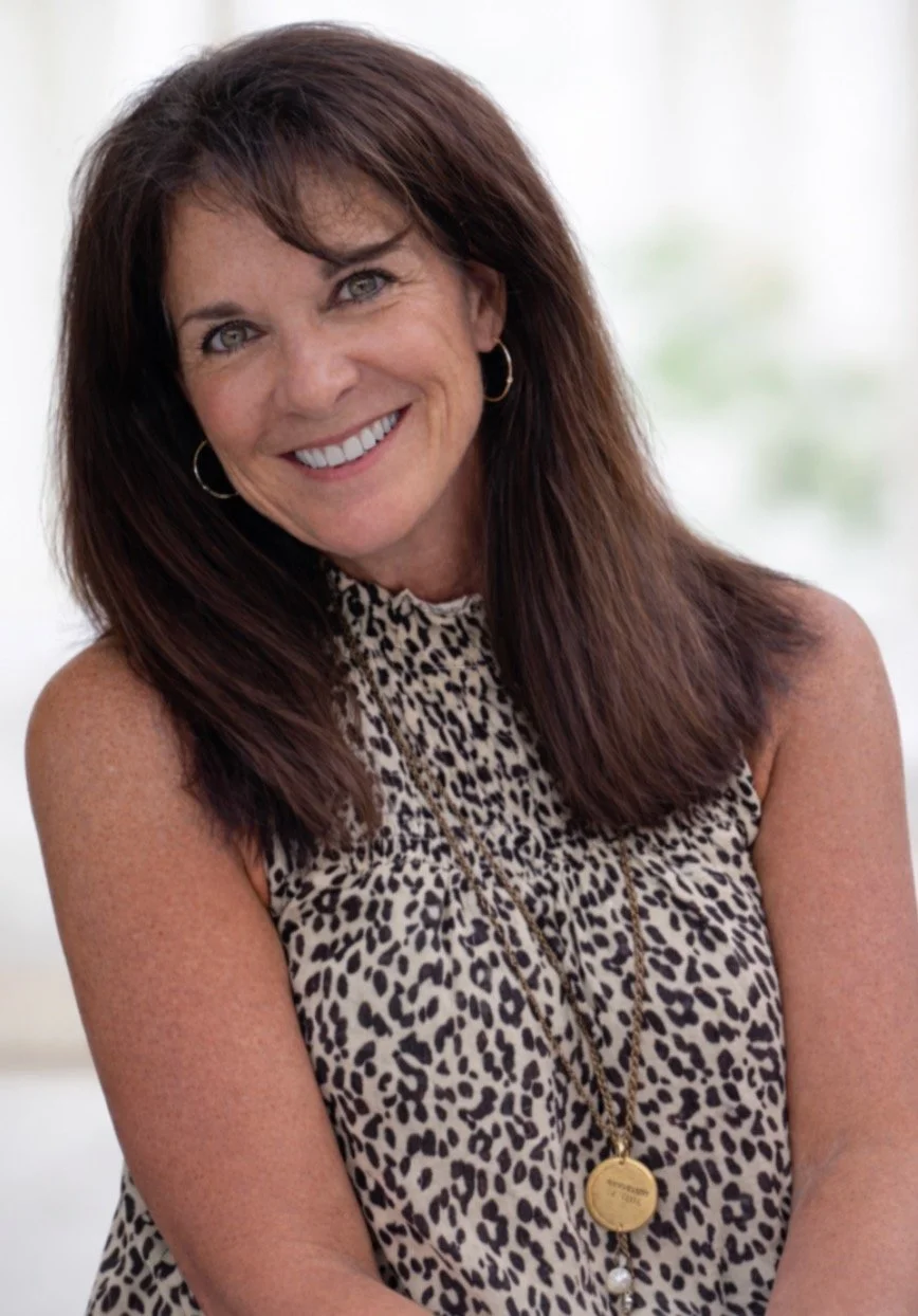 A woman with shoulder-length brown hair smiling, wearing hoop earrings, a leopard print sleeveless top, and a gold necklace with a round pendant.