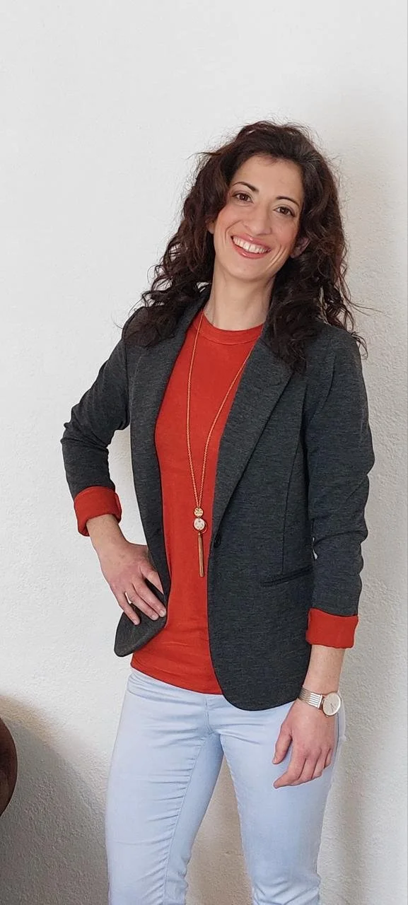 A woman with curly brown hair smiling, wearing a dark blazer over a red top, white pants, a long necklace, and a watch, standing against a plain white wall.