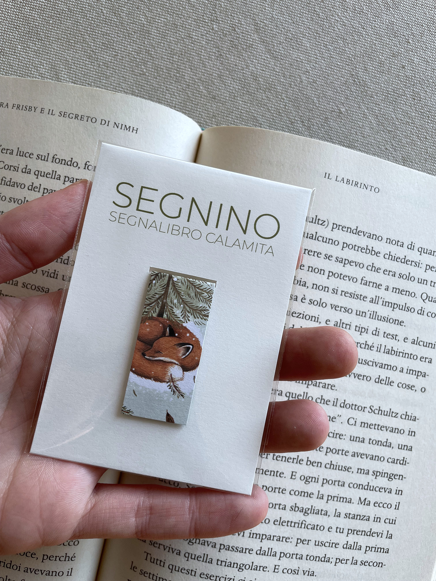 Segnalibro artigianale magnetico chiamato 'Segnino', realizzato con scarti di lavorazione. Nella confezione si vede il segnalibro illustrato con una volpe addormentata sotto rami di pino. Una mano lo tiene in primo piano sopra le pagine aperte di un