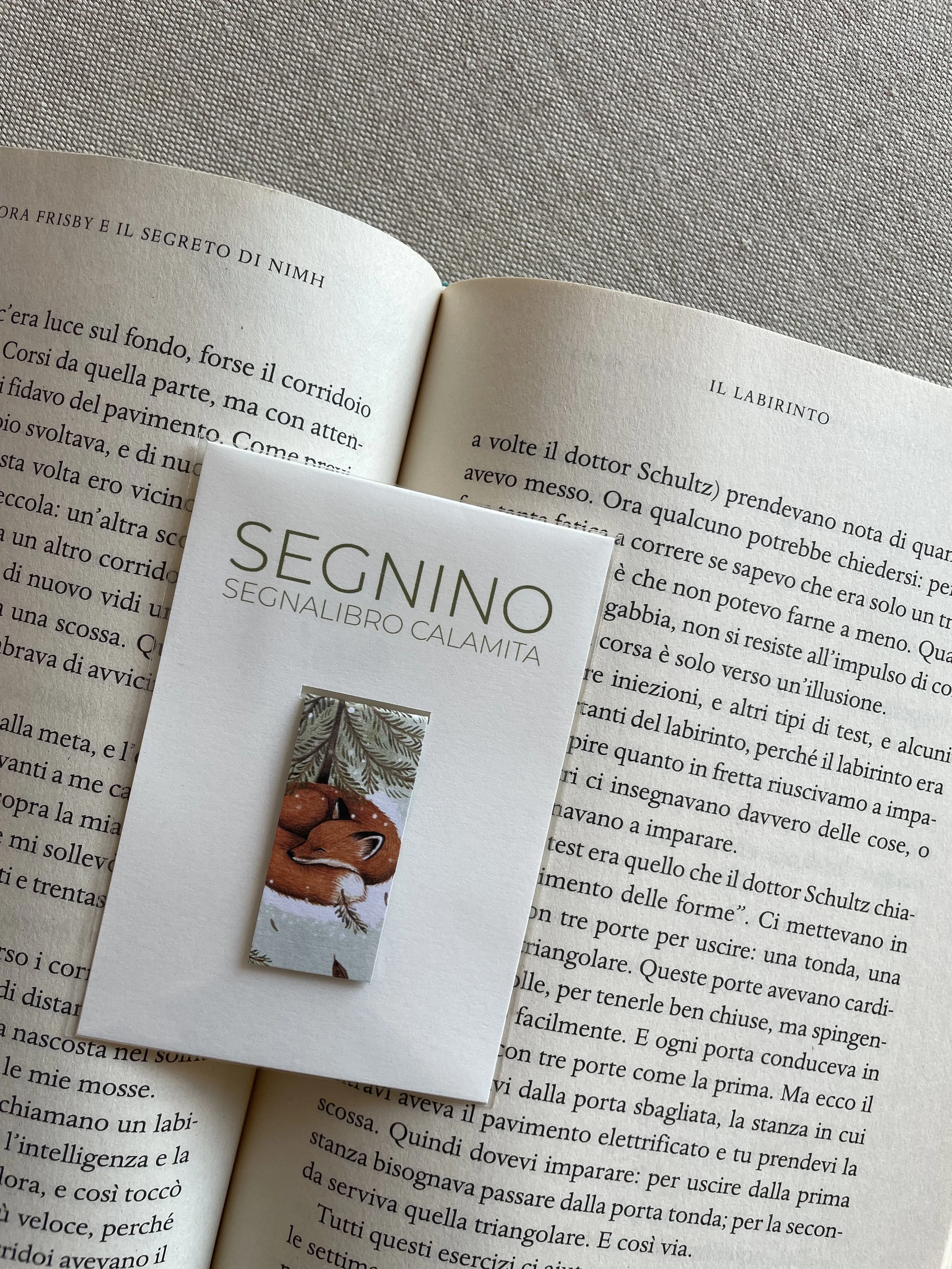 Segnalibro artigianale magnetico chiamato 'Segnino', realizzato con scarti di lavorazione. Nella confezione si vede il segnalibro illustrato con una volpe addormentata sotto rami di pino. è appoggiato tra le pagine di un libro