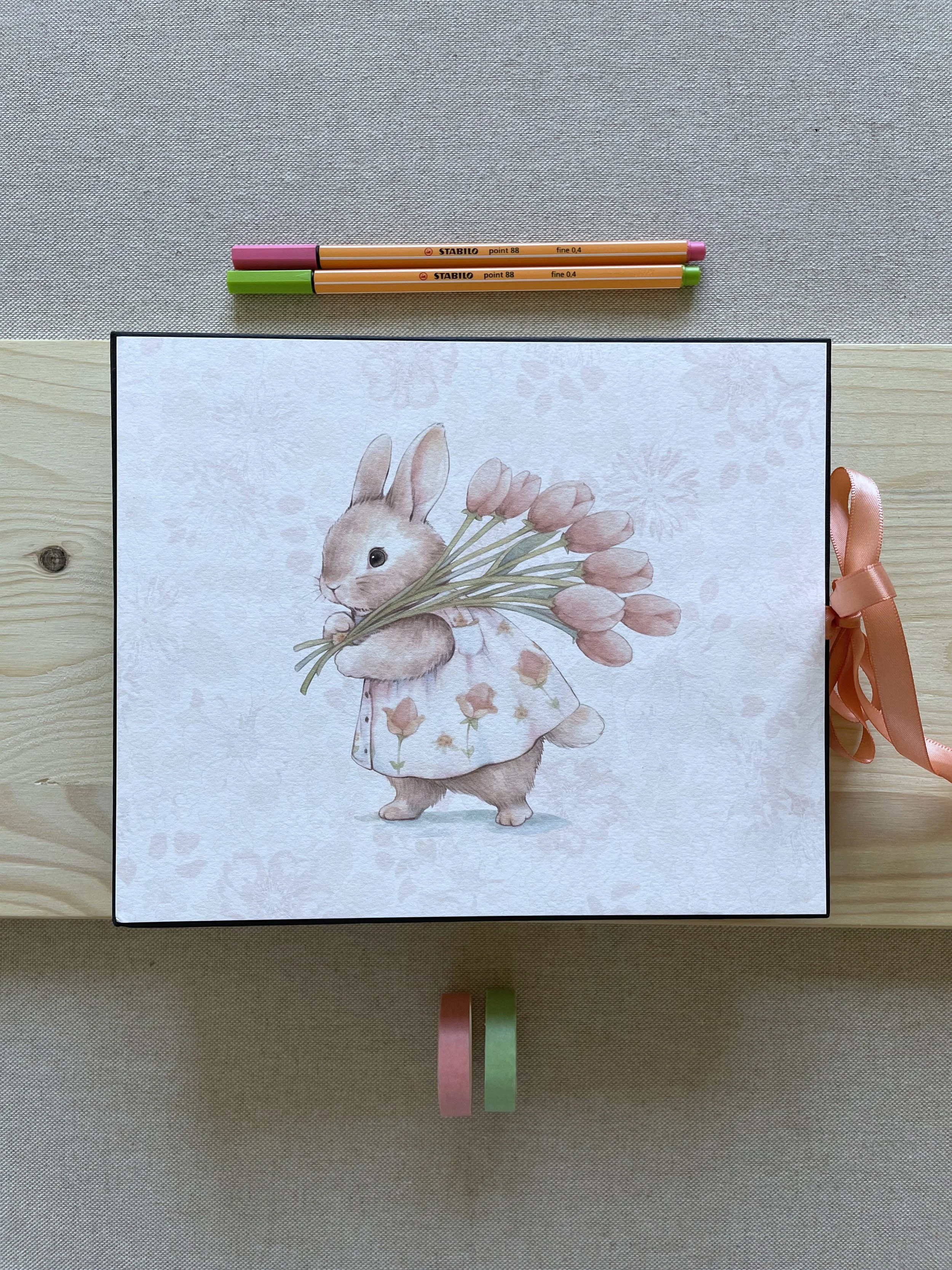 Album per foto ad anelli  con copertina decorata con un coniglietto che tiene un mazzo di tulipani, su un tavolo. Il kit comprende anche due penne colorate e due nastrini colorati per incollare le foto. realizzato artigianalmente.