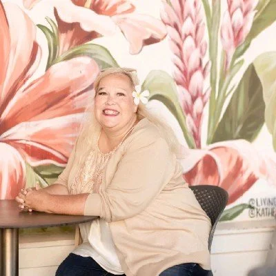Woman sitting at a table with a floral mural background, smiling with a flower in her hair.