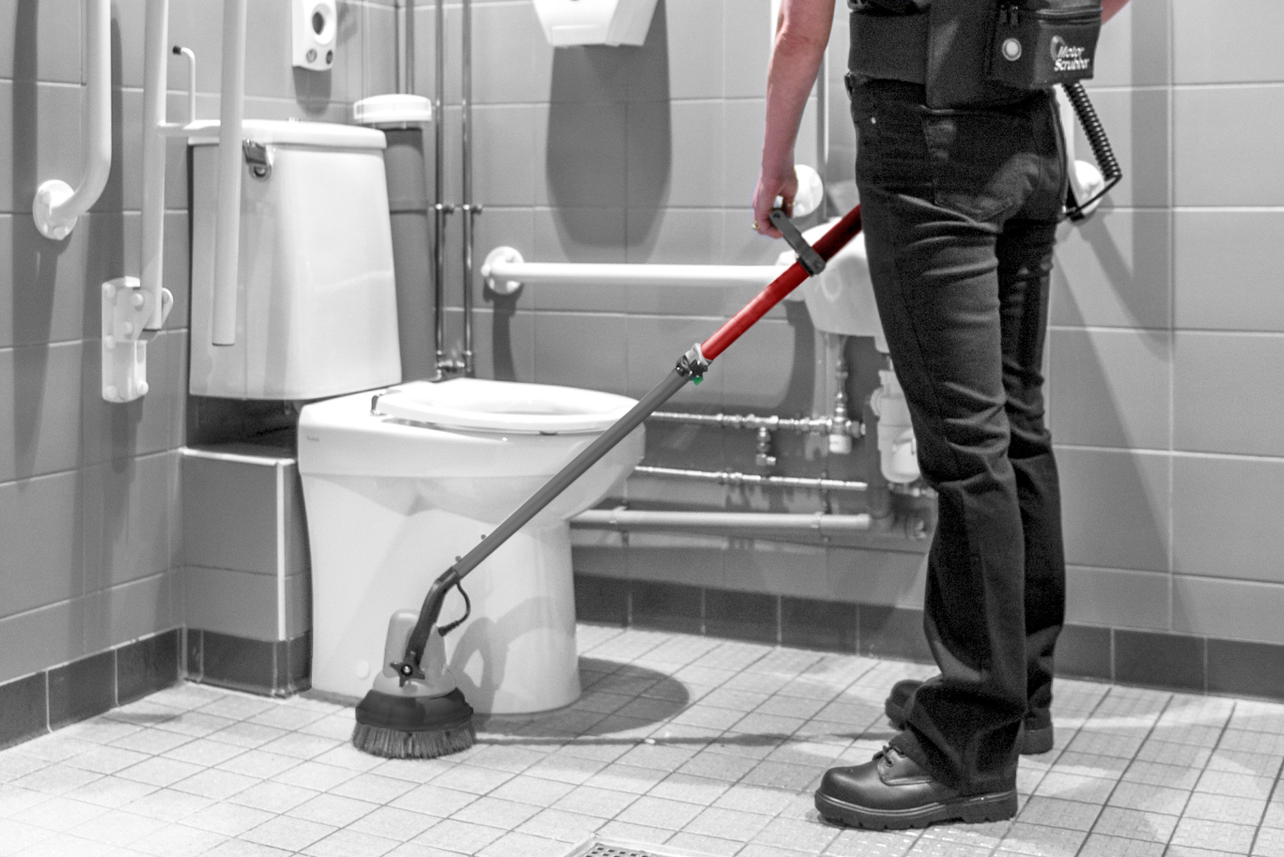 Operative using the MotorScrubber M3 with extended handle and backpack battery to clean floor tiles around a toilet in a commercial washroom, demonstrating access to confined and hard-to-reach areas.