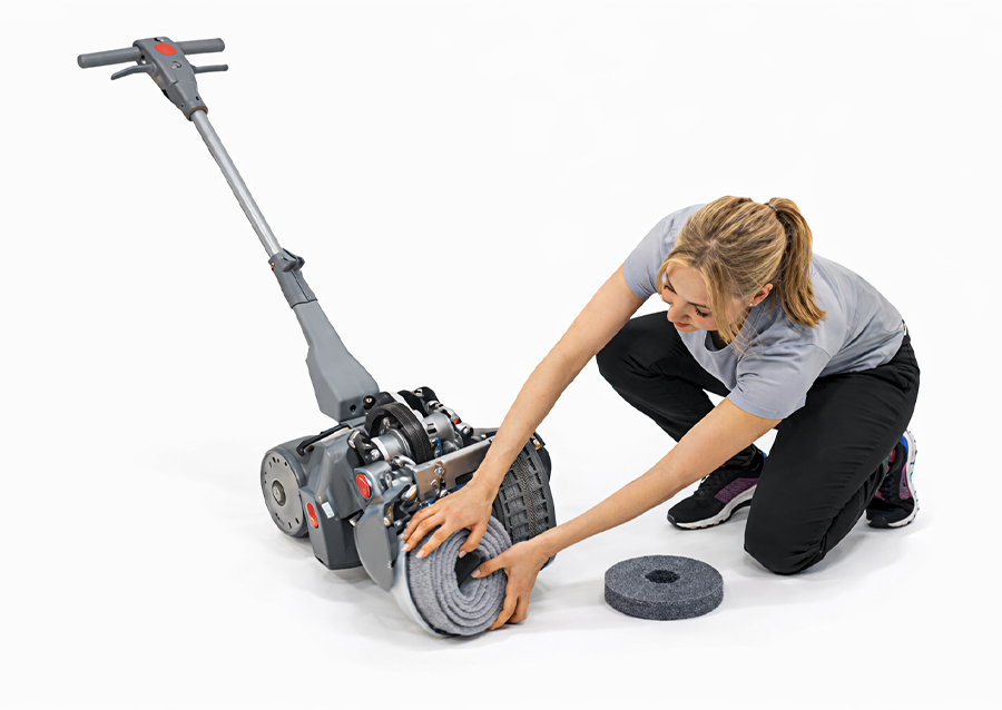 Operator kneeling to change a traditional rotary brush on a walk-behind scrubber dryer, showing a low, awkward pull-and-twist changeover that can slow teams down and add strain.