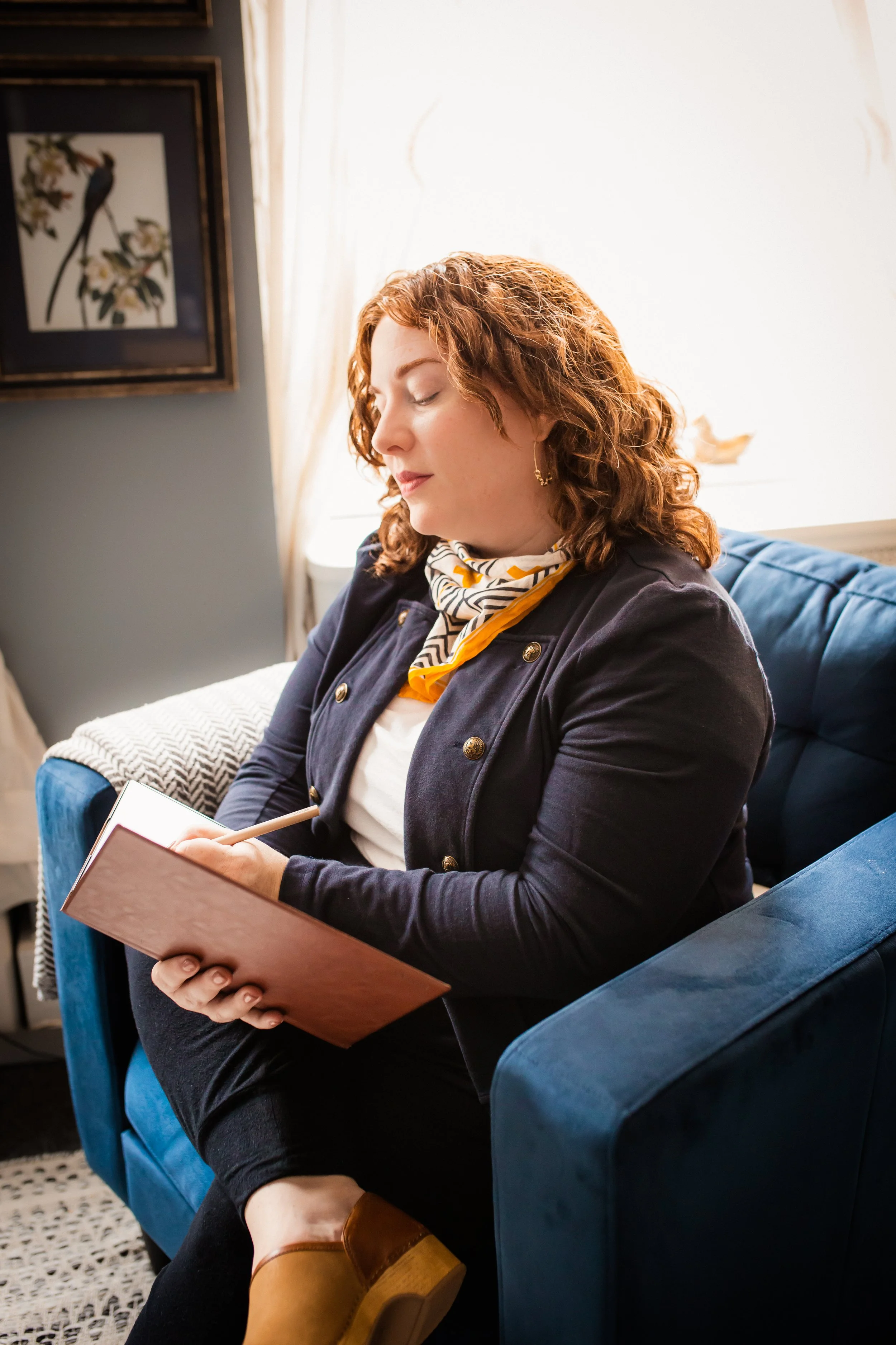 A woman with curly red hair and a scarf, sitting on a blue couch, writing in a notebook.