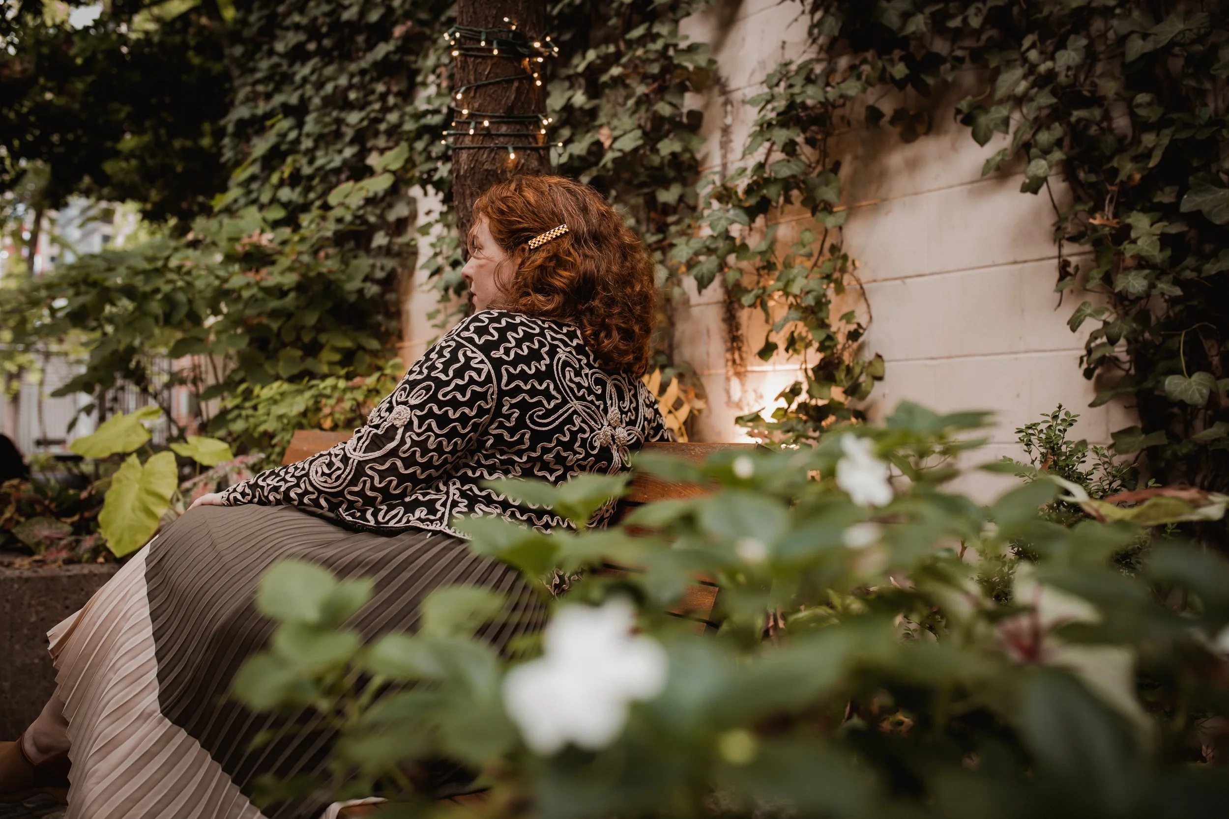 A woman with curly hair wearing a graphic black and white top sitting on a wooden bench in a garden with green foliage and white flowers nearby, under string lights hanging from a tree.