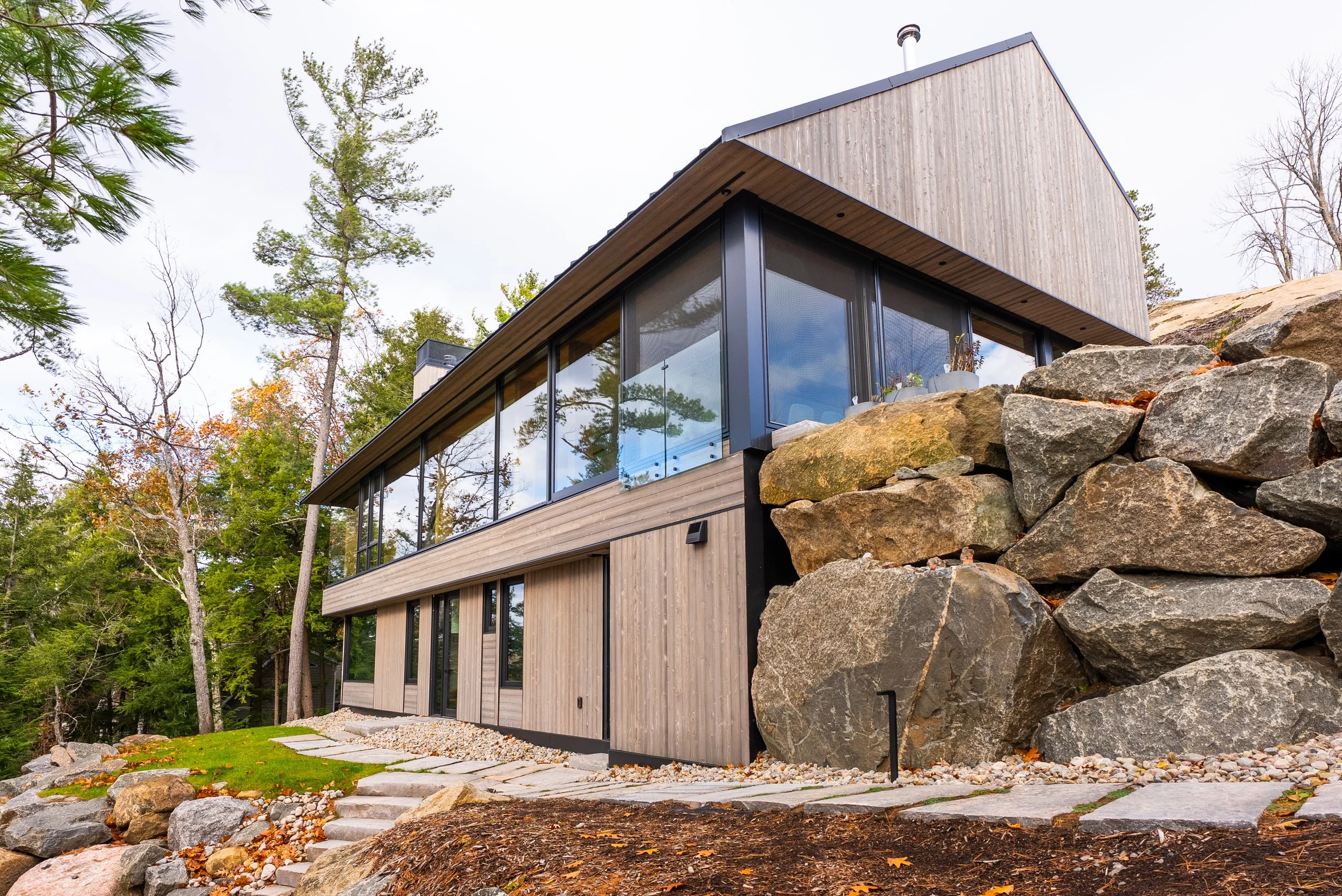 Modern house with large glass windows, wooden exterior, and surrounded by rocks and trees.