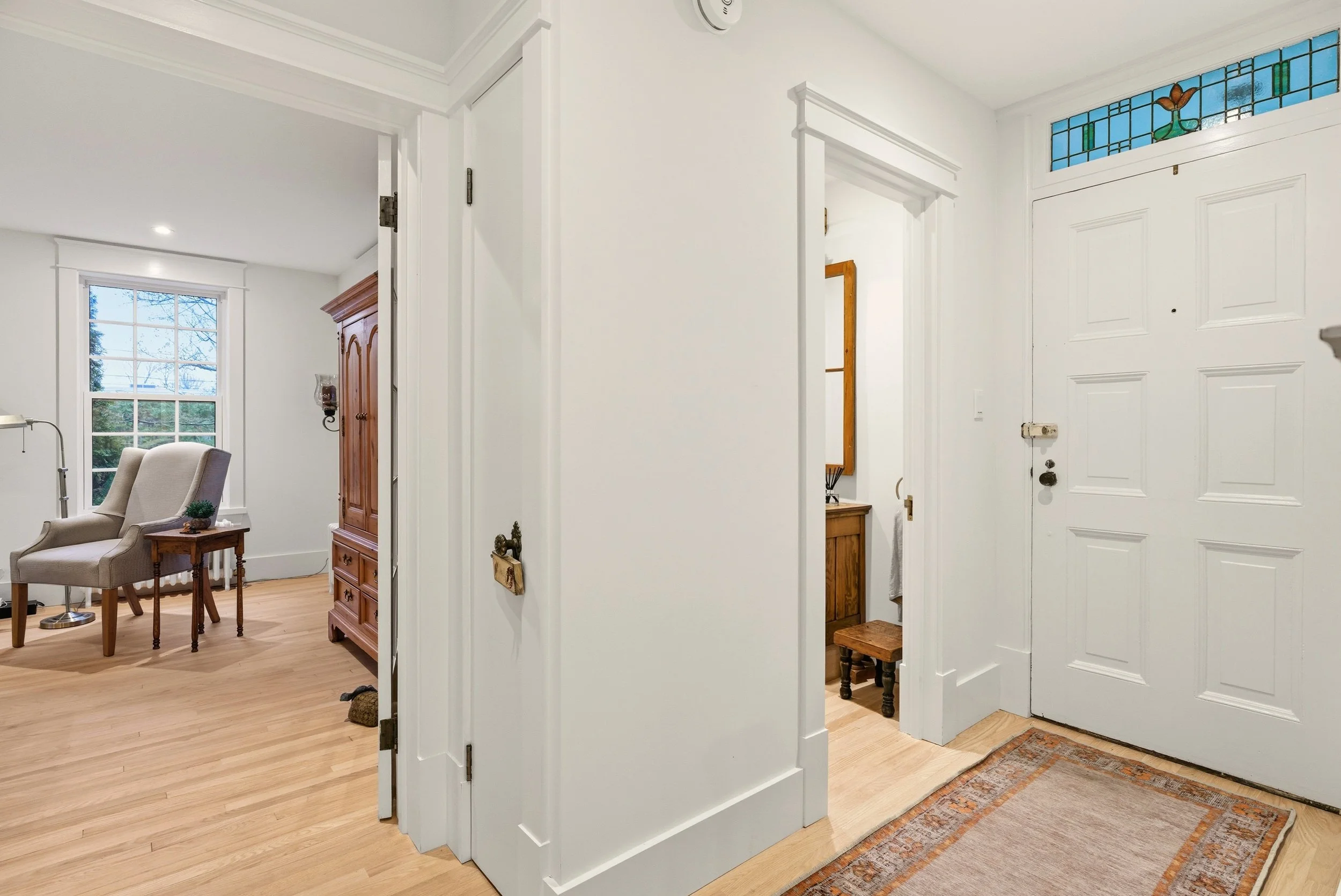 Entryway with white door, stained glass window, beige rug, and two small rooms with wooden furniture, hardwood floors, and white walls.