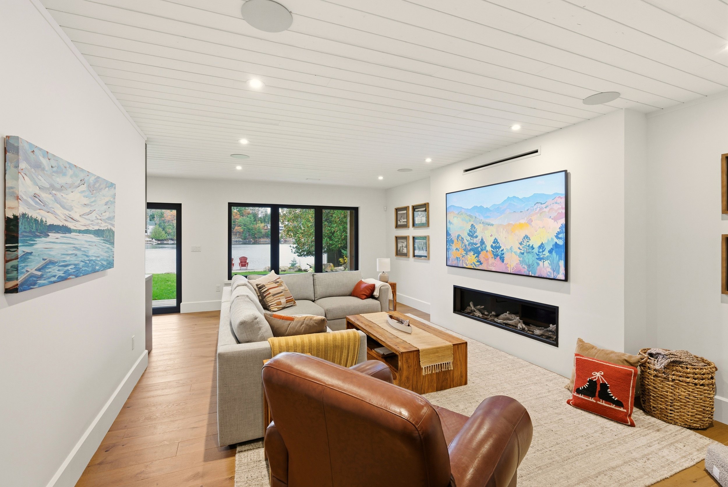 Living room with large window overlooking a lake, featuring a beige sectional sofa, a wooden coffee table with a striped table runner, a leather armchair, a painting of a mountain and lake scene on the wall, a large photo display on the opposite wall