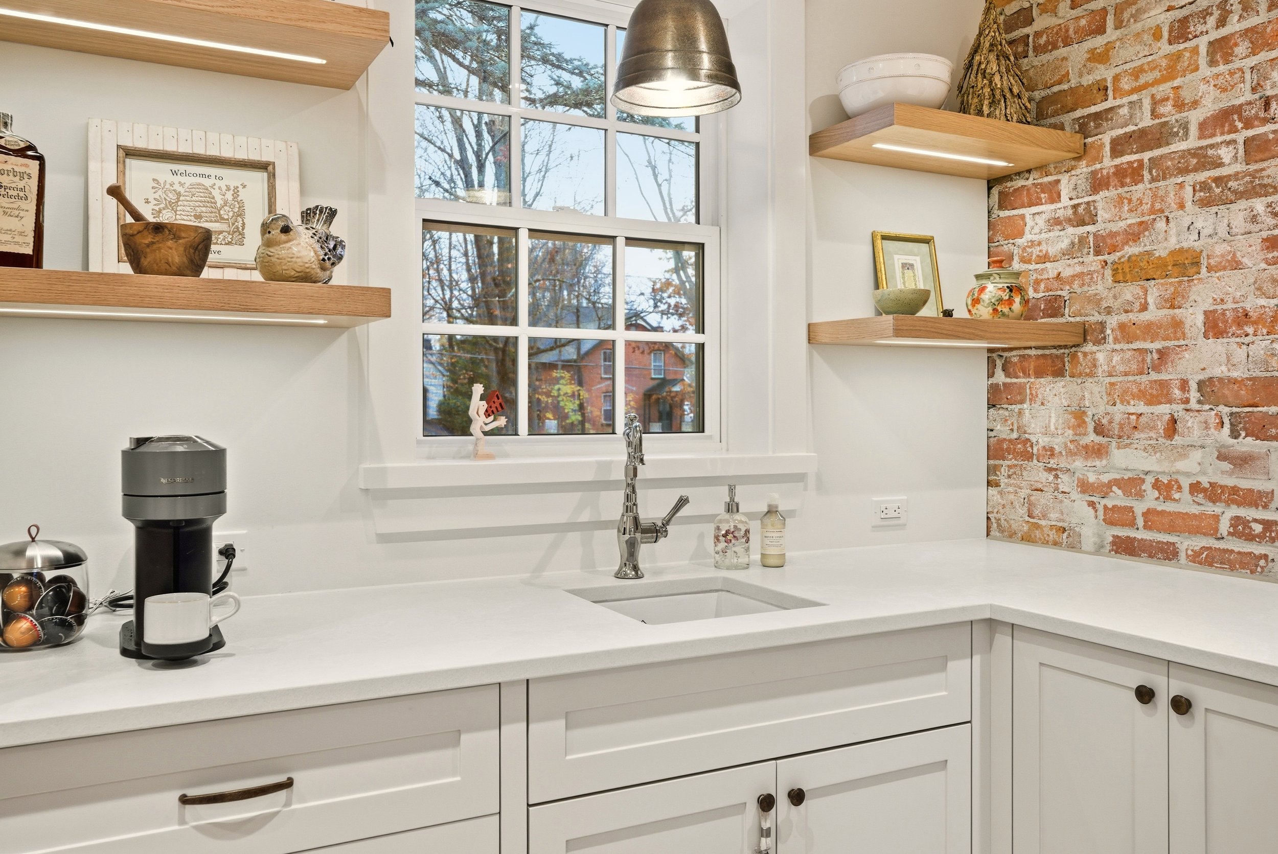 Cozy kitchen with white cabinetry, a brick accent wall, open wooden shelves with decorative items, a window showing outdoor trees, stainless steel faucet, coffee machine, and soap dispensers.