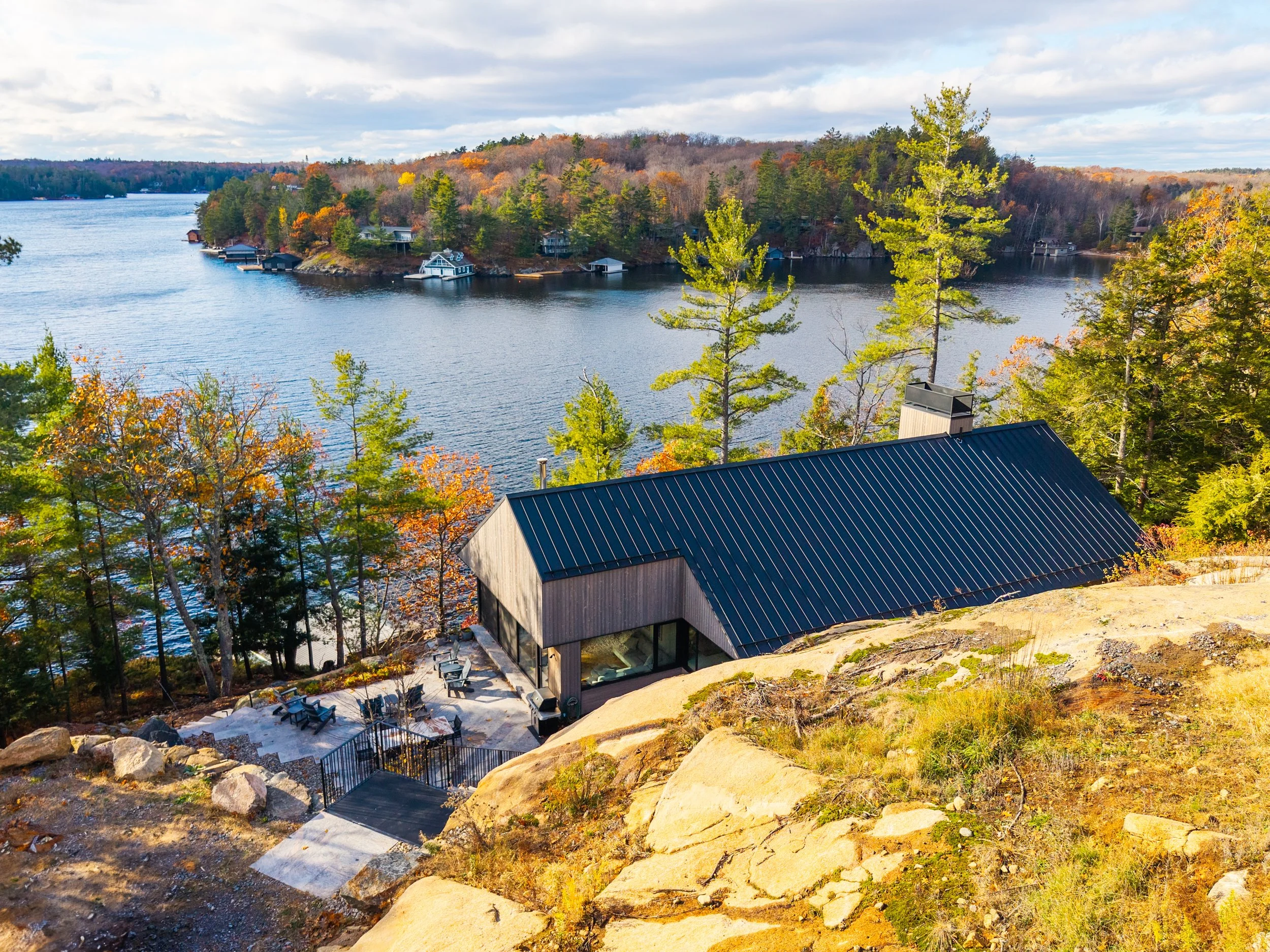 A modern house with a black metal roof nestled among trees on a hillside overlooking a lake with small boats and houses along the shoreline, during autumn.
