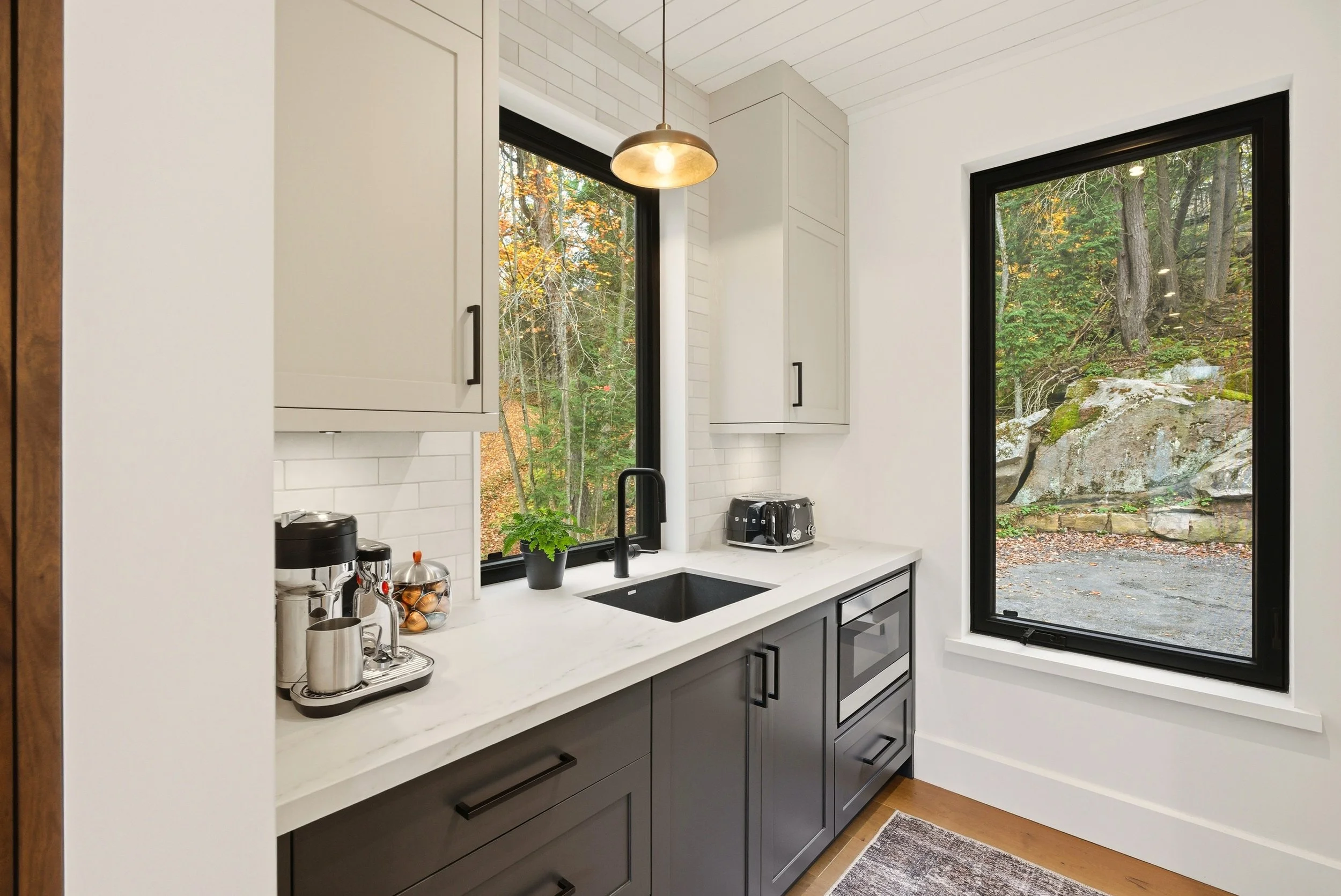 Modern kitchen with white walls, gray lower cabinets, and black handles. There is a black sink, a window with a view of trees and rocks, and small kitchen appliances like a toaster and coffee maker on the white countertop. A potted plant is near the 