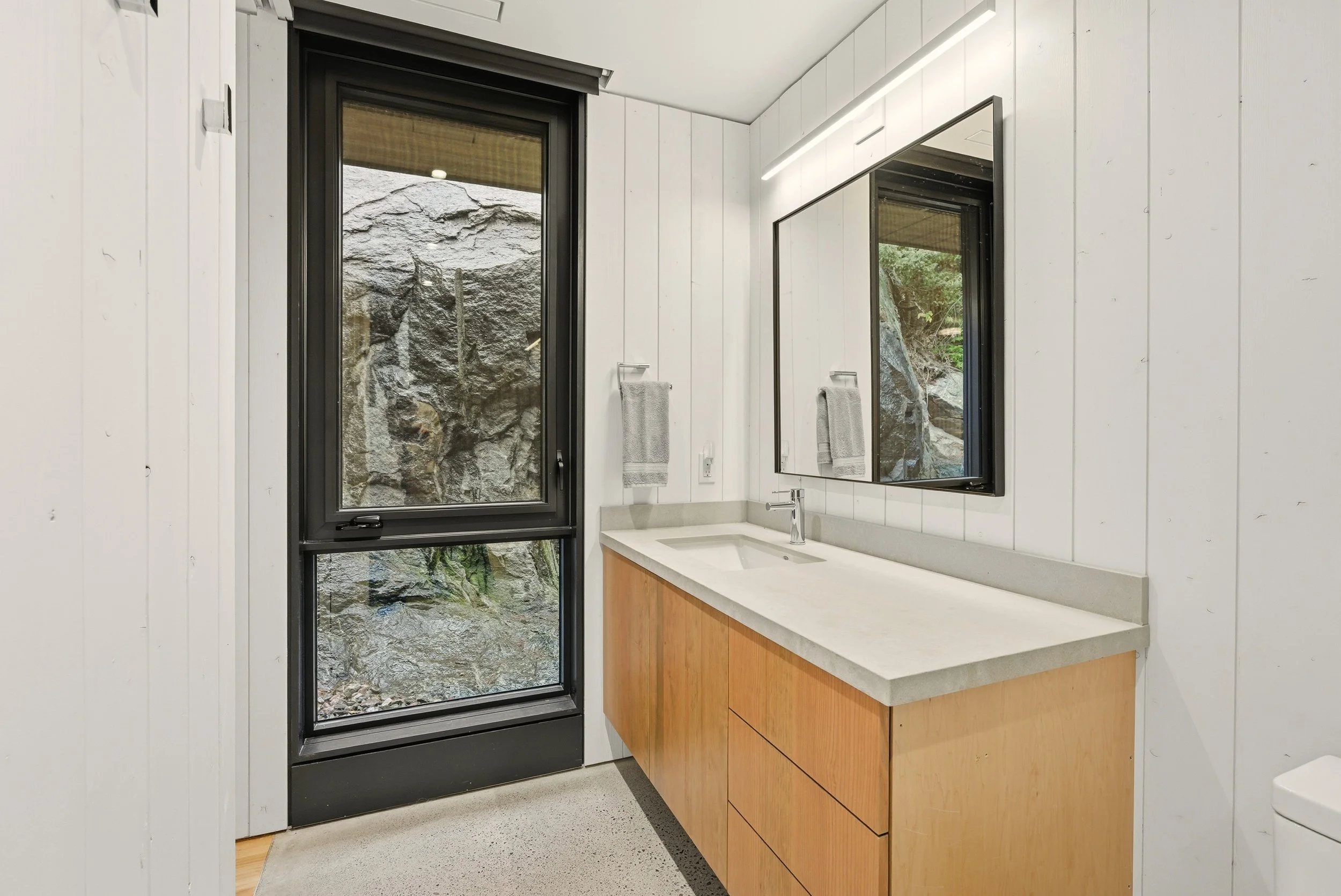 Modern bathroom with a white wooden wall, a large mirror, a beige countertop with a sink, two gray towels on the walls, a window with a rocky outdoor view, and a white toilet partially visible on the right.