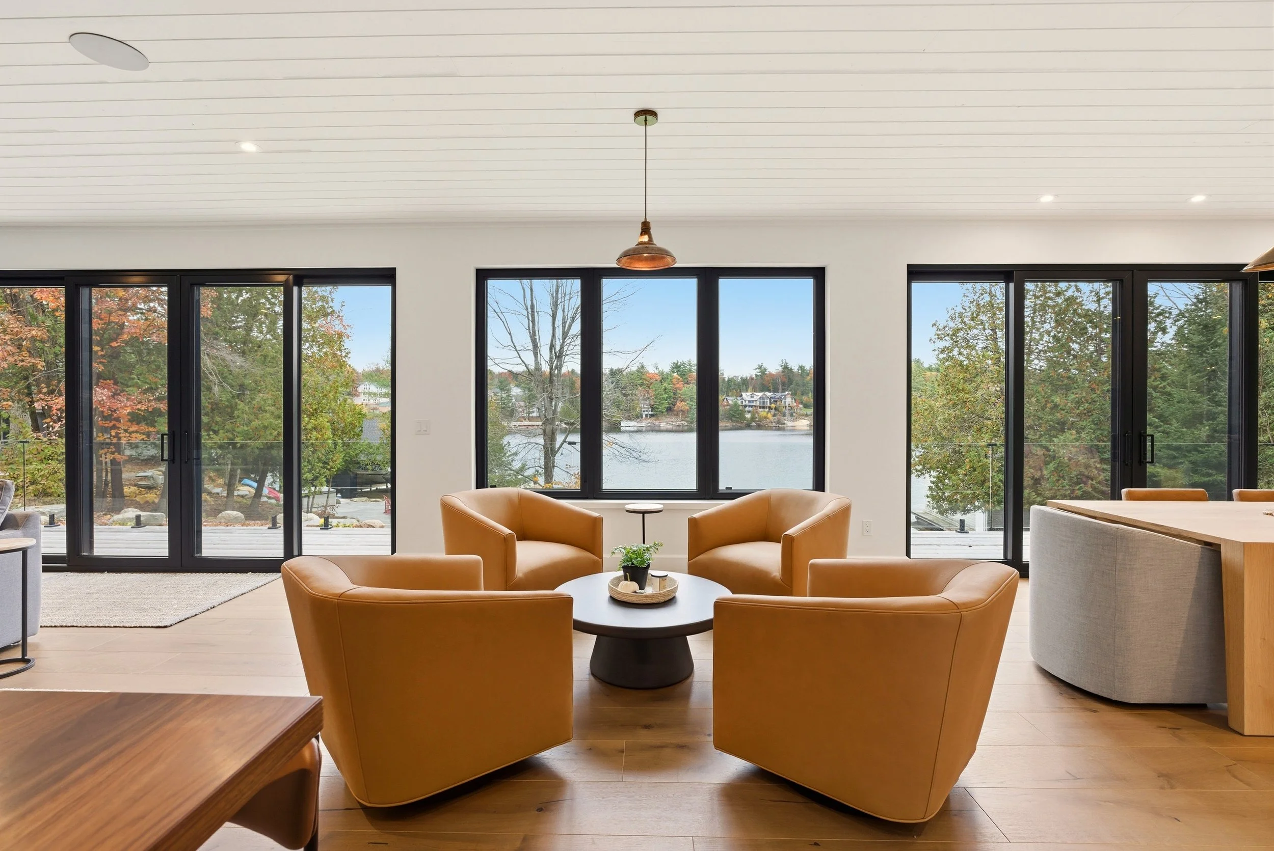 Modern living room with four tan leather armchairs arranged around a black coffee table, large windows showing an outdoor lake view, and a white ceiling with recessed lighting and a pendant light.