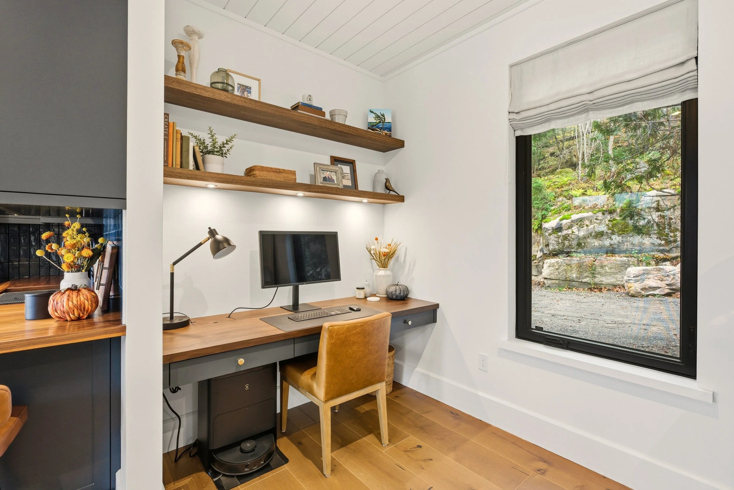 Home office with a wooden desk, a computer monitor, a yellow chair, and two wooden floating shelves with decorative items, next to a large window overlooking a rocky, wooded landscape.