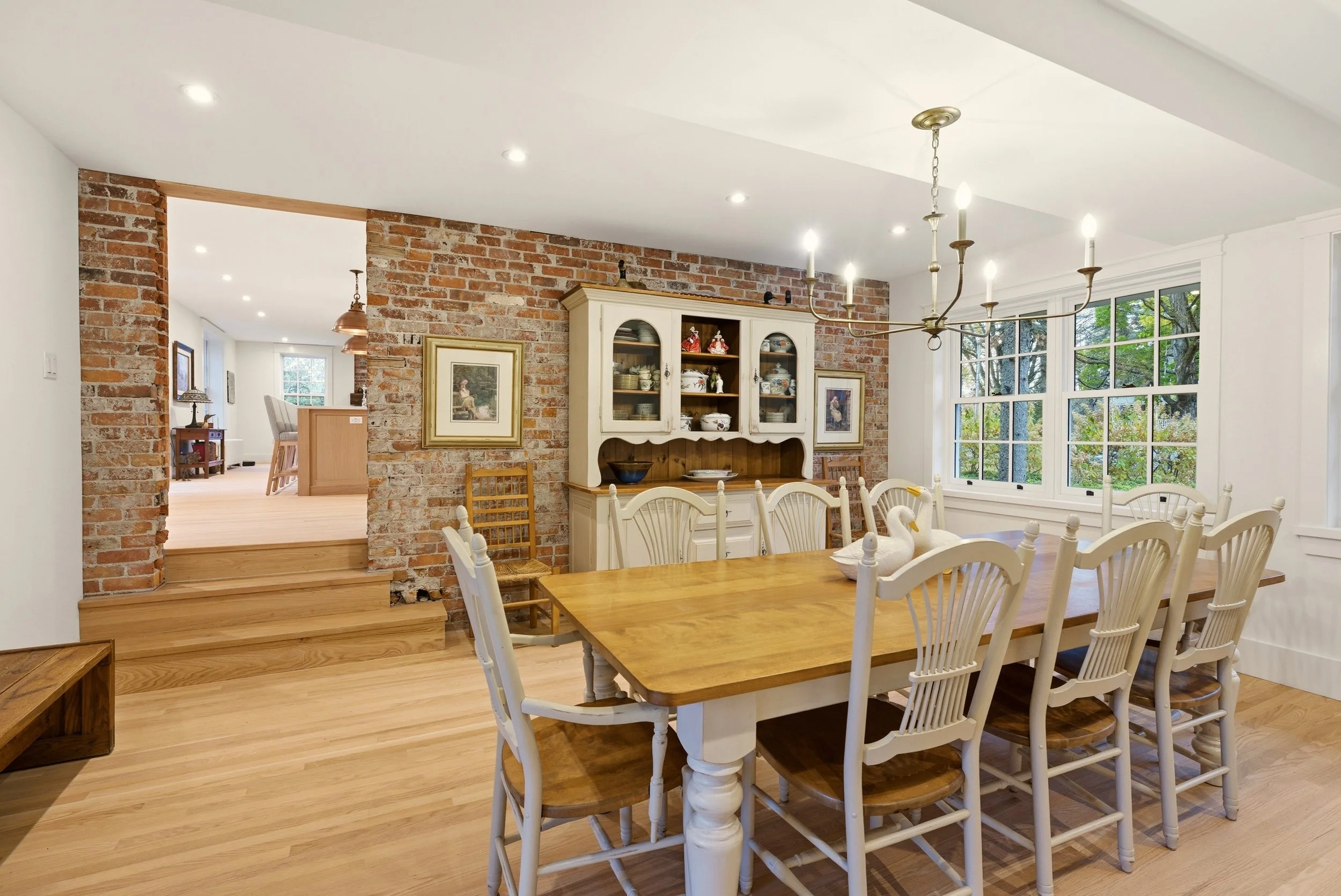 A dining room with a wooden table surrounded by white chairs, a white hutch against a brick accent wall, a chandelier overhead, and large windows showing greenery outside.