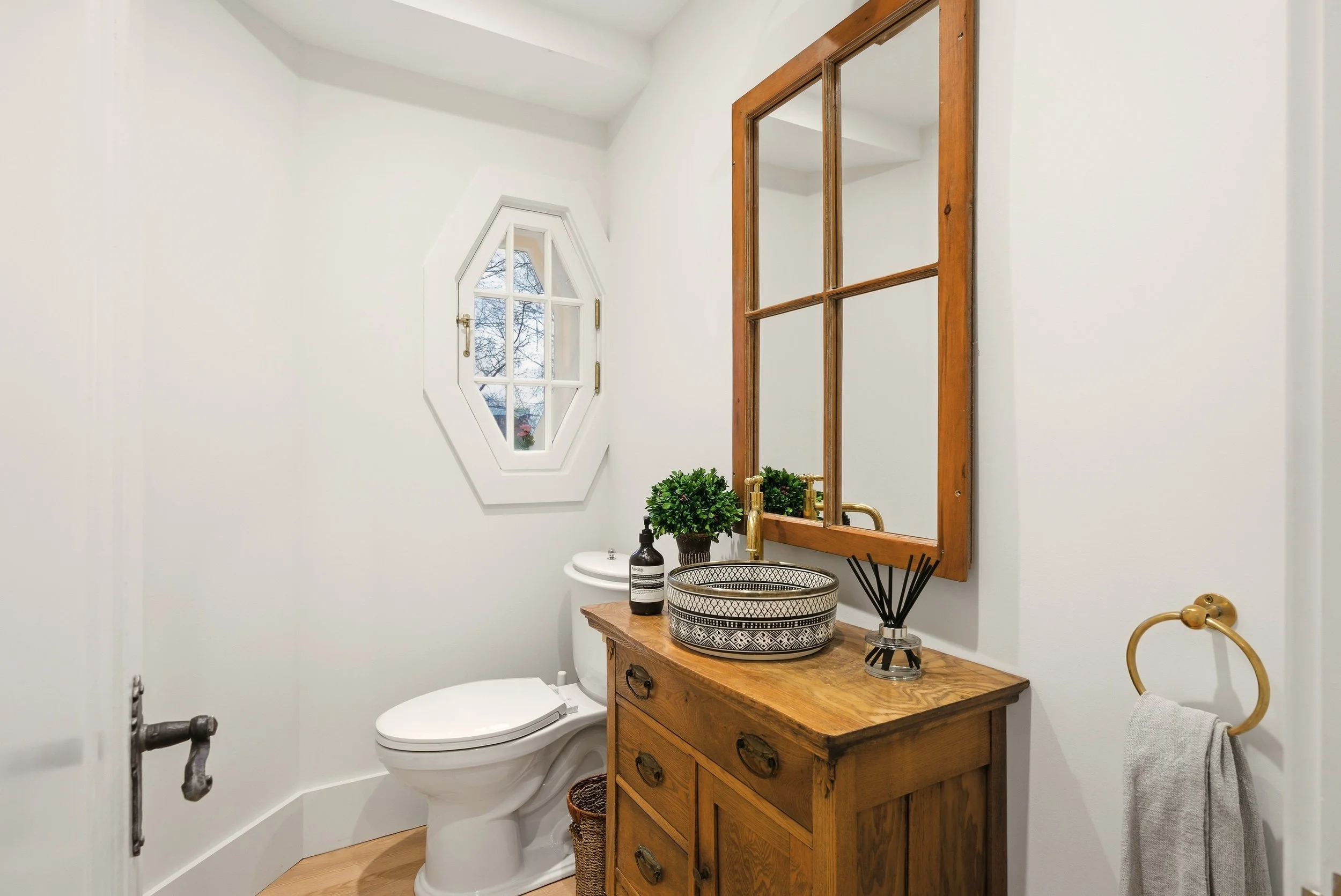 A small bathroom with white walls, a toilet, wooden vanity with a black and white patterned basin sink, a wooden framed mirror, a small window with white trim, a potted plant, a bottle of soap, a reed diffuser, and a towel ring with a towel.