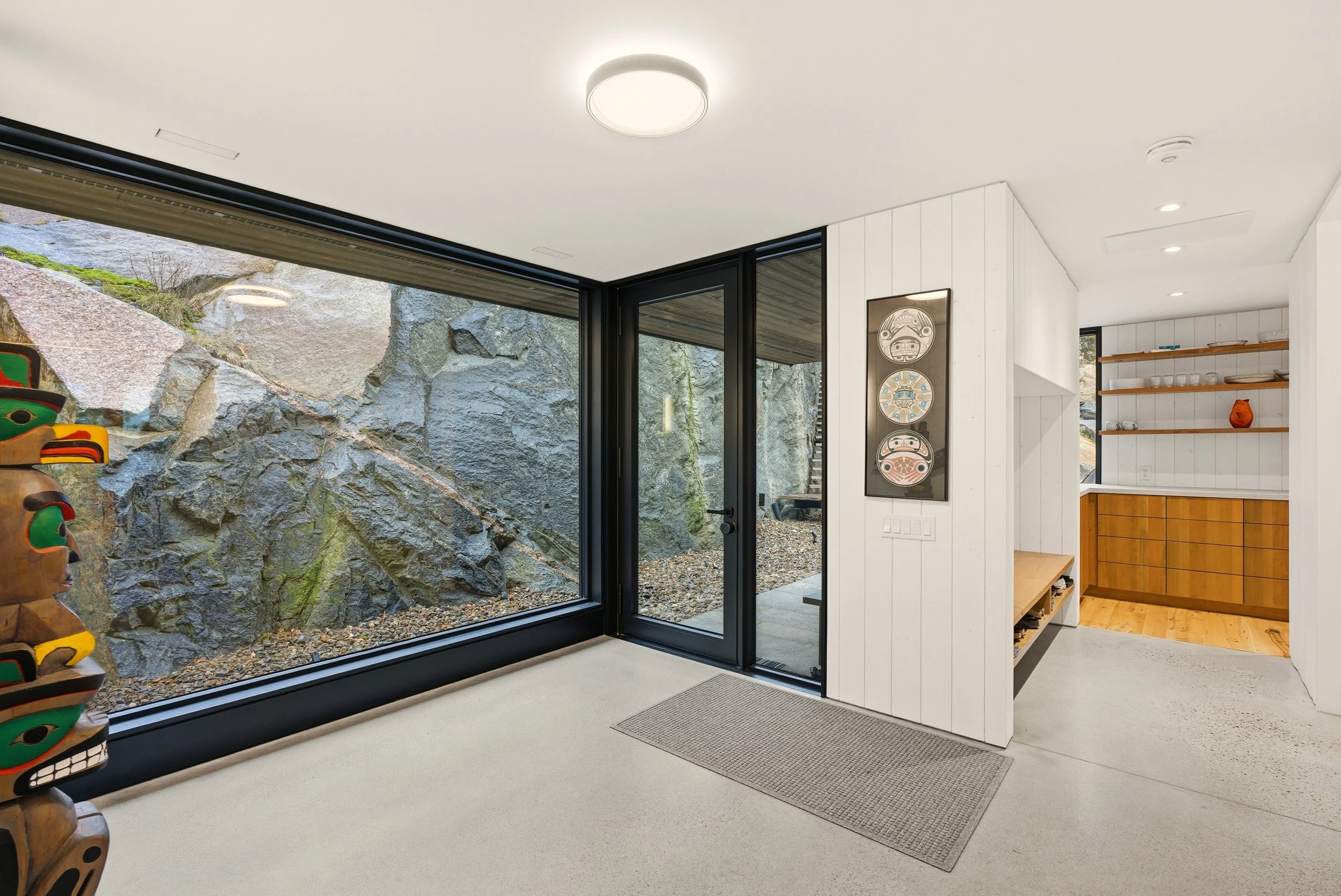 Interior view of a modern house with large glass windows showing a rocky outdoor terrain, white walls, wooden shelves with dishes, and a corner with colorful tribal art on the left.