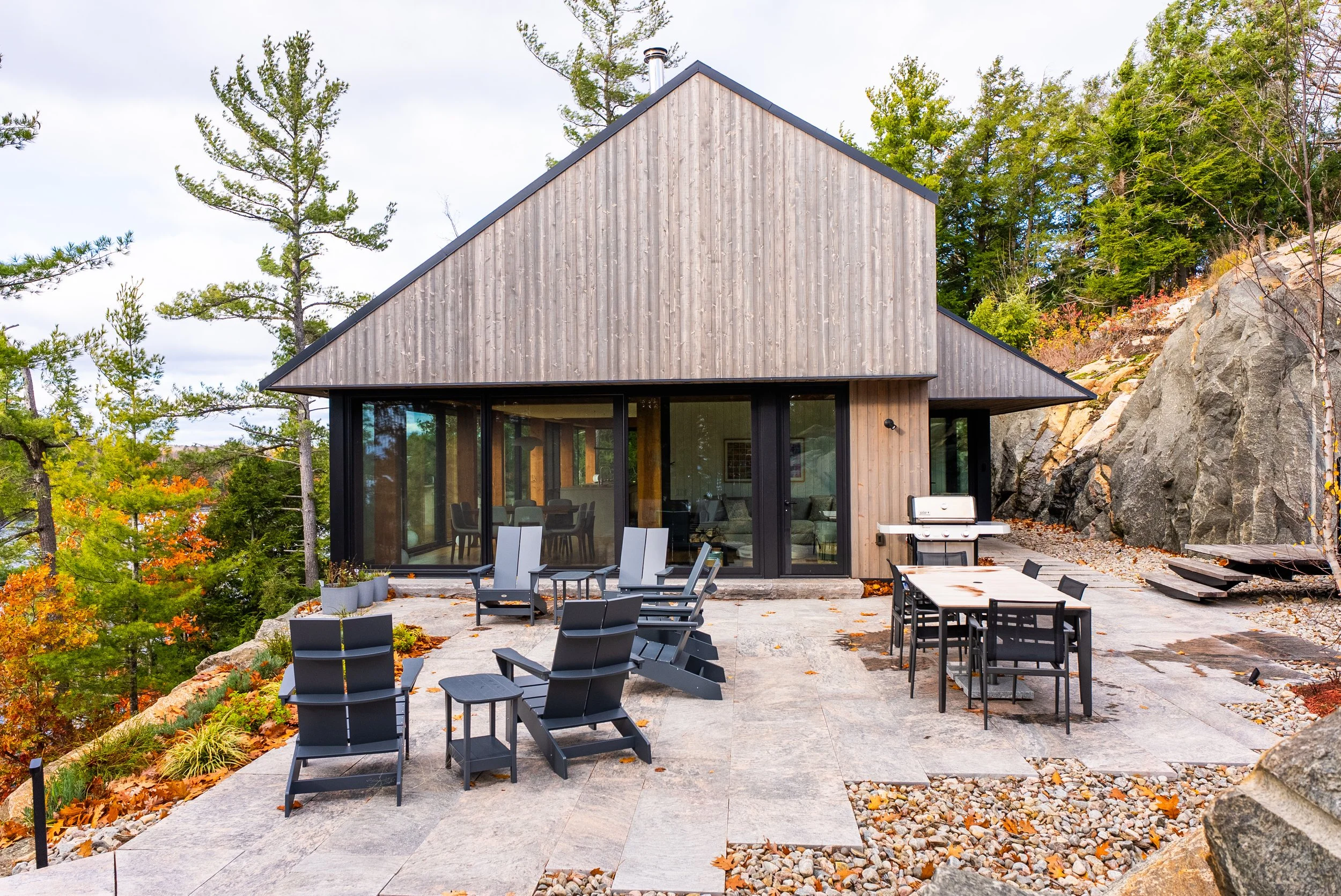 Modern house with large glass windows, outdoor seating area with chairs and tables, grill, and surrounded by autumn trees and rocks.