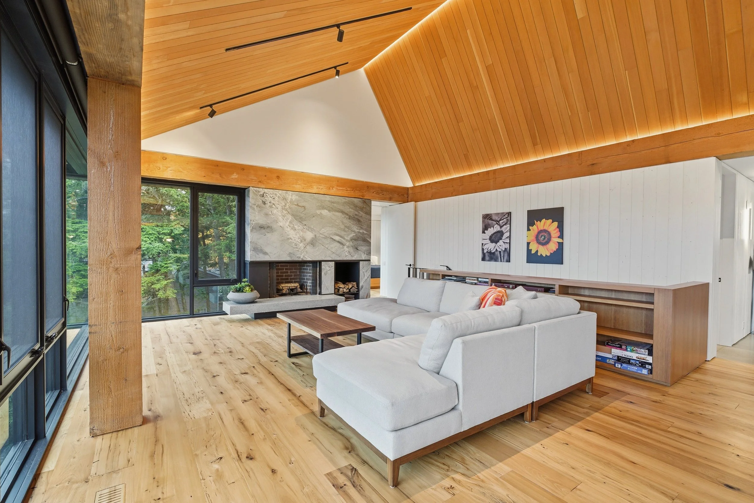 Living room with wooden floors and ceiling, white sectional sofa, fireplace with marble surround, large windows showing green trees outside, wood trim, artwork of flowers on wall, and a coffee table.