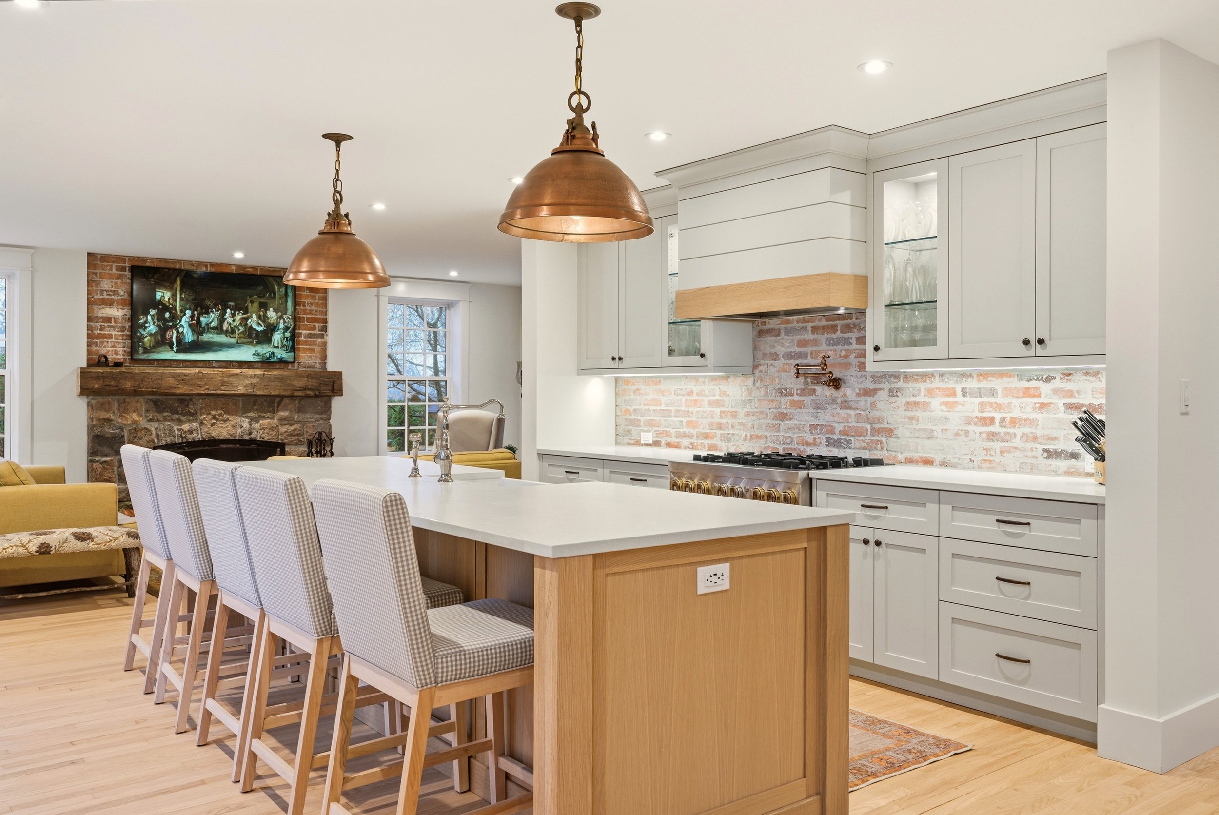 Modern kitchen with white cabinets, brick backsplash, and a kitchen island with a white countertop and checkered chairs. Hanging copper pendant lights over the island. In the background, a living area with a stone fireplace and a TV.