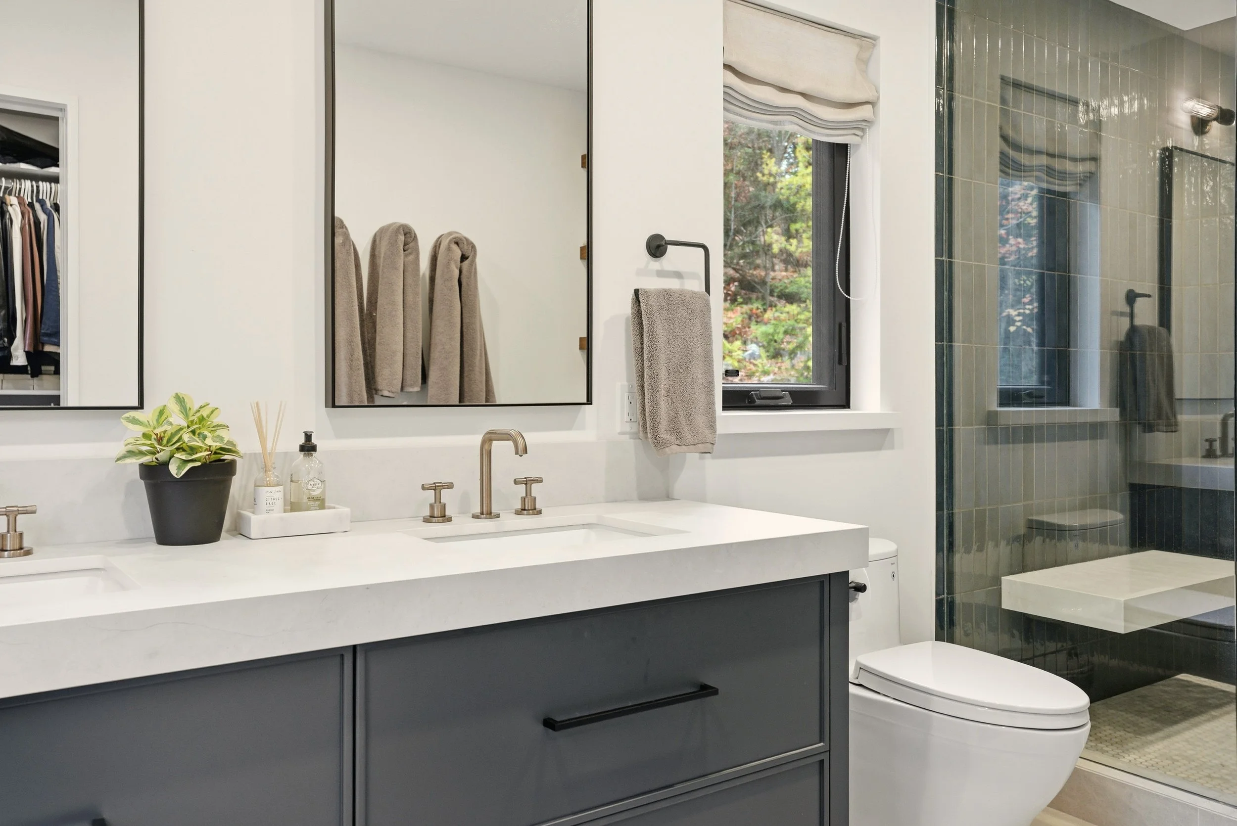 Modern bathroom with double vanity, large mirrors, a plant, toiletries, a towel, window with outside greenery, and a walk-in shower with glass enclosure.