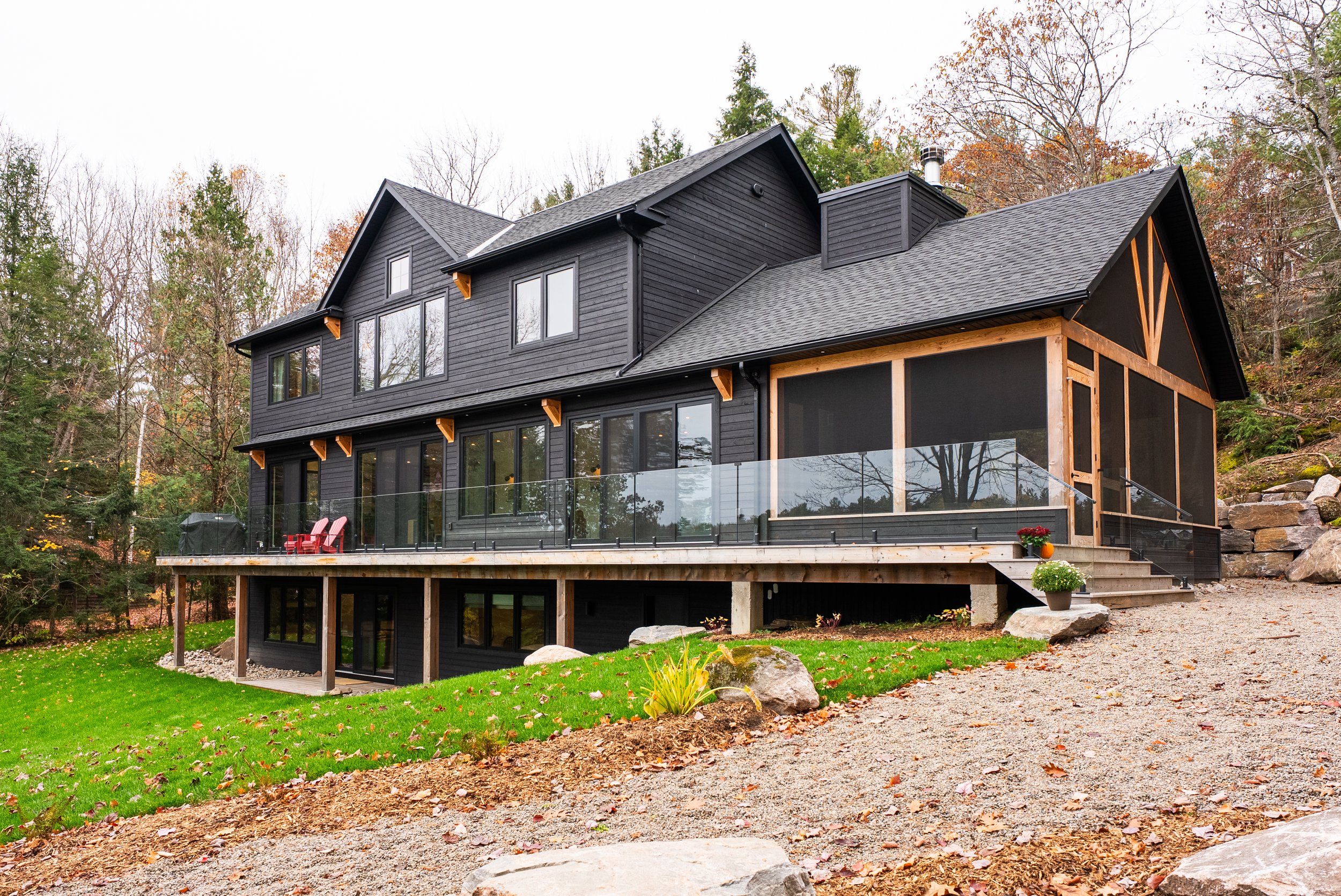 A modern black wooden house with a large wraparound deck, glass railing, and large windows, situated in a wooded area with fall trees in the background.