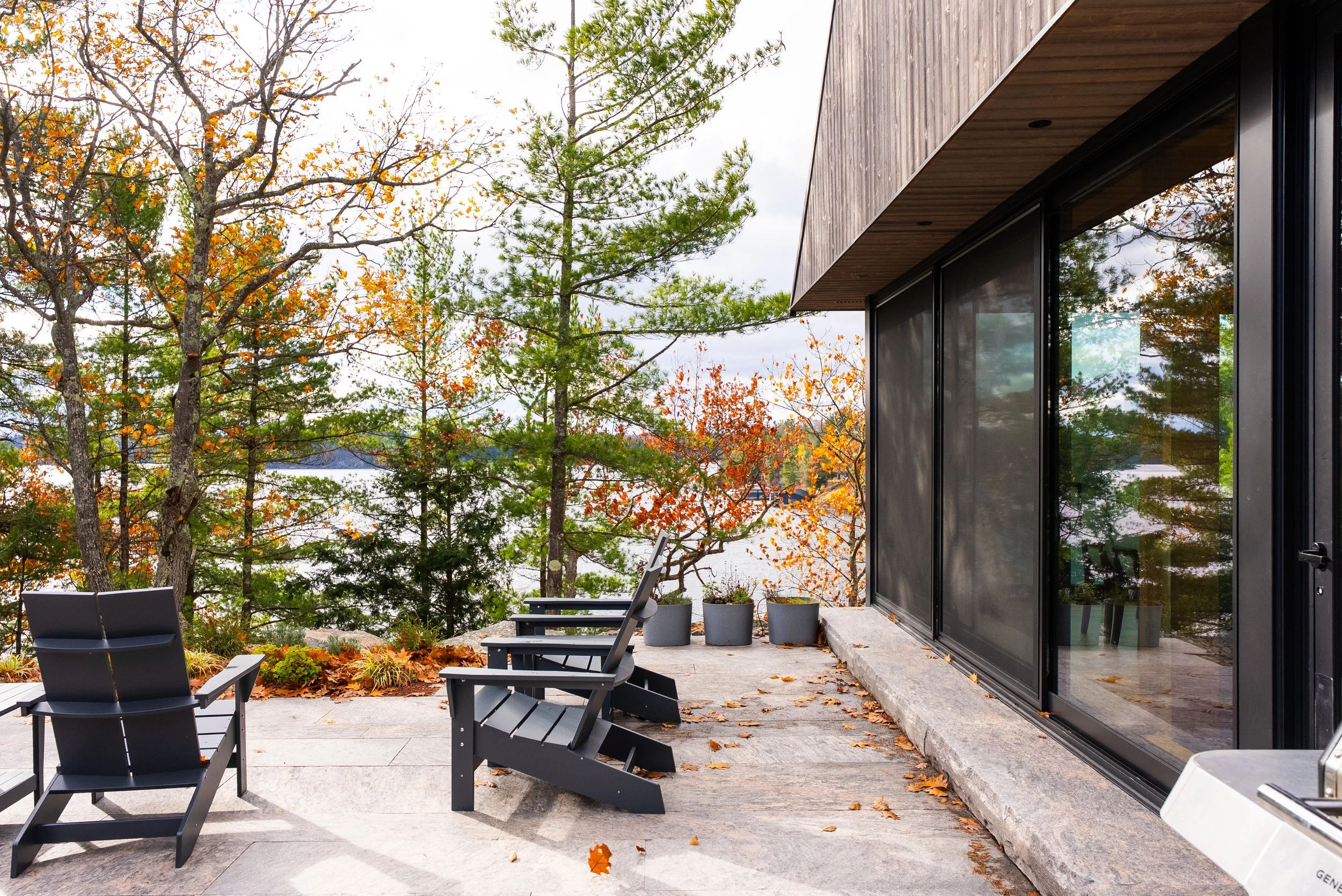 Outdoor patio with black Adirondack chairs and potted plants, overlooking a lake surrounded by trees with autumn foliage.
