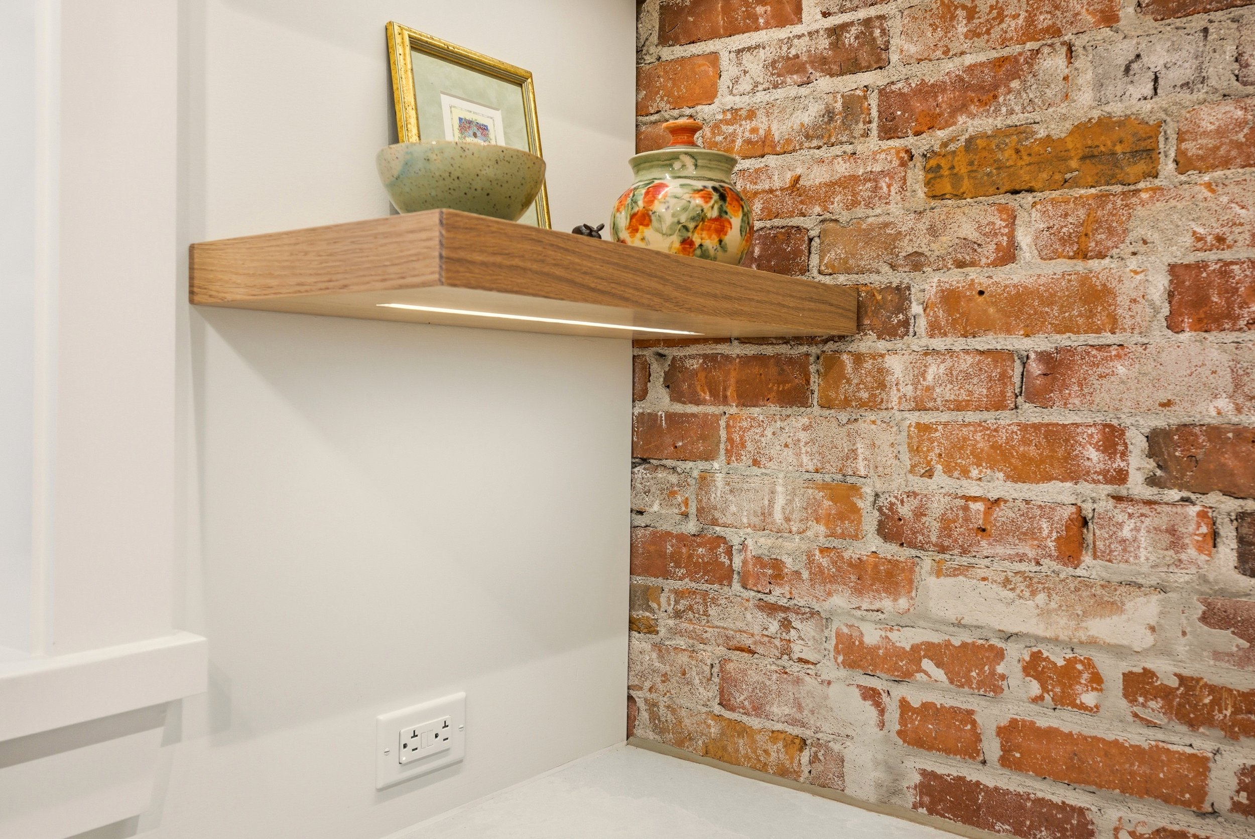 A corner with a brick wall, a wooden floating shelf holding decorative pottery and a framed picture, a white wall with an electrical outlet, and a white countertop.