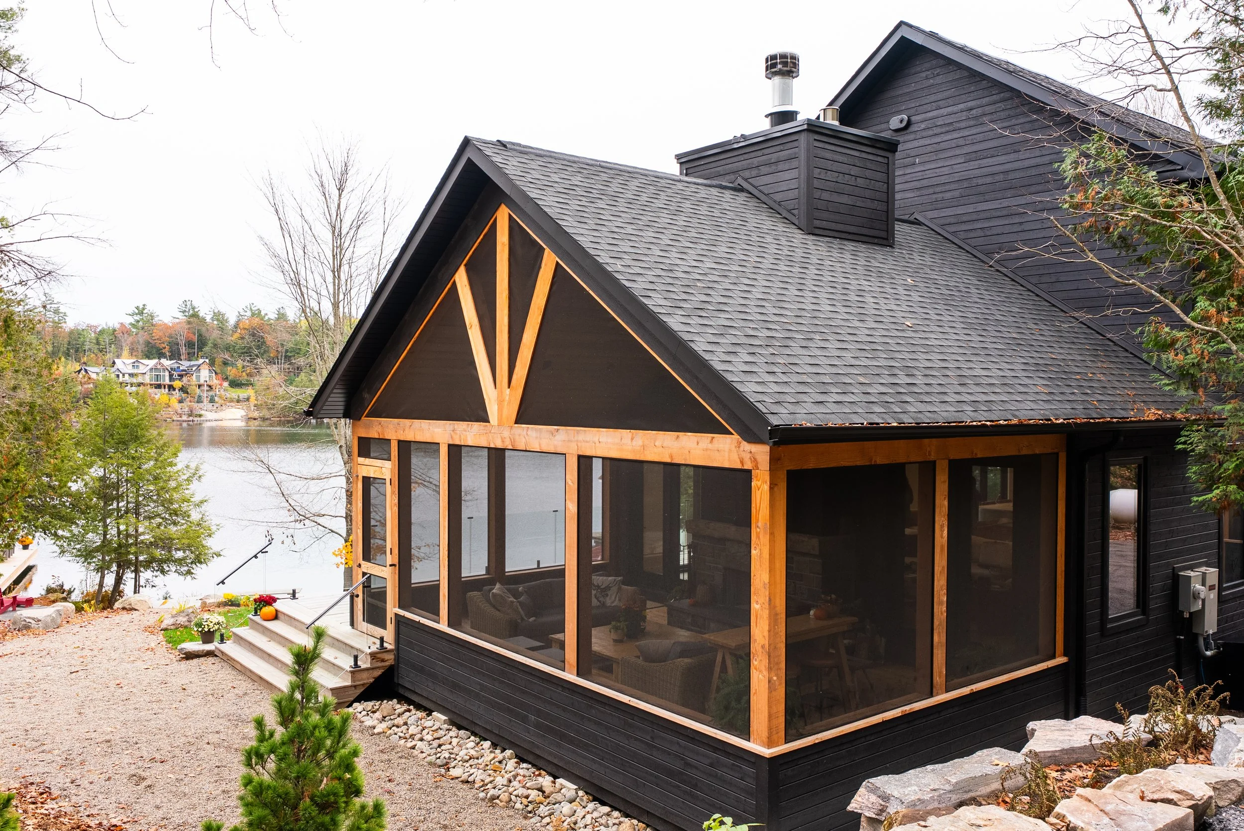 Black house with a screened porch, situated near a lake, surrounded by trees, with a staircase leading up to the porch decorated with pumpkins and flowers, and a rocky landscaped area in the foreground.