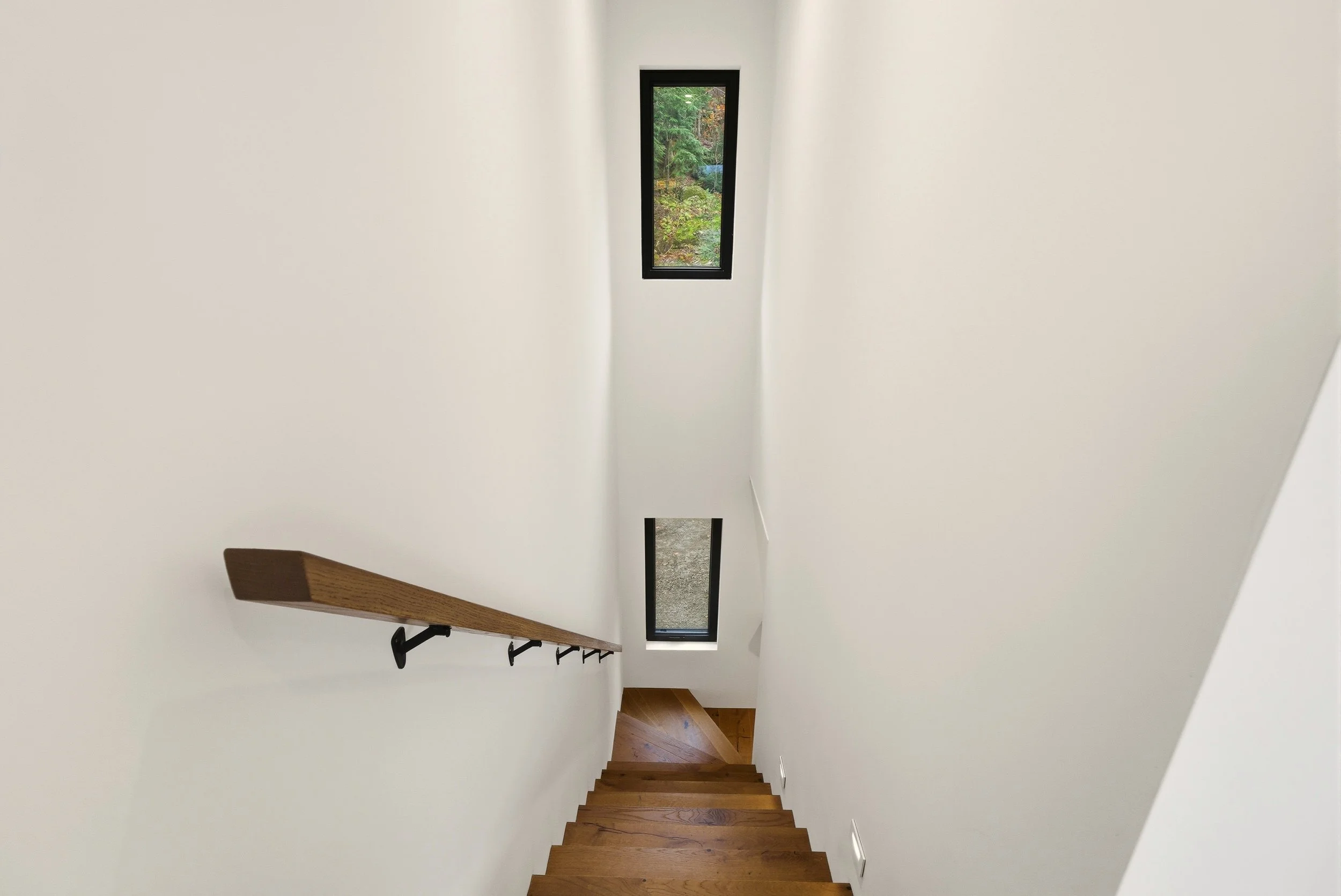 View of a staircase from the top, with wooden steps, a dark brown handrail on the left, and two narrow vertical windows with views of greenery, walls painted white.