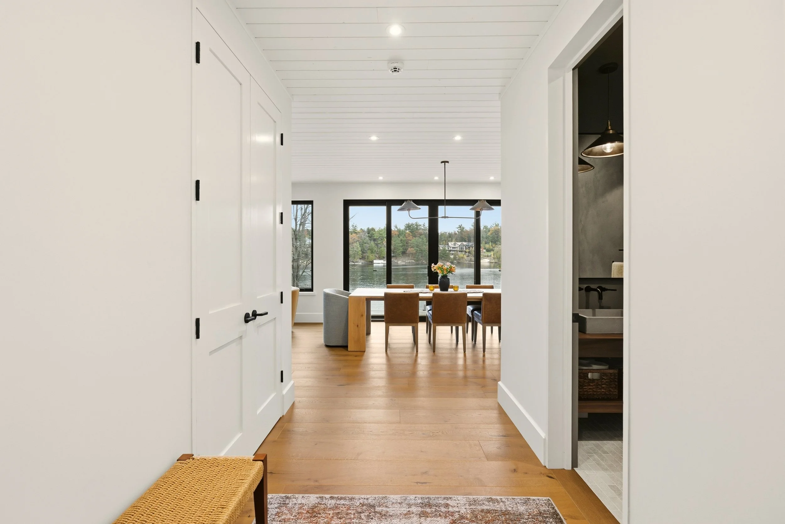 View of a bright dining area with a large window overlooking a lake, wooden dining table with six chairs, and pendant lights hanging from a white ceiling. Part of an adjacent room with a small sink and dark wall is visible on the right.