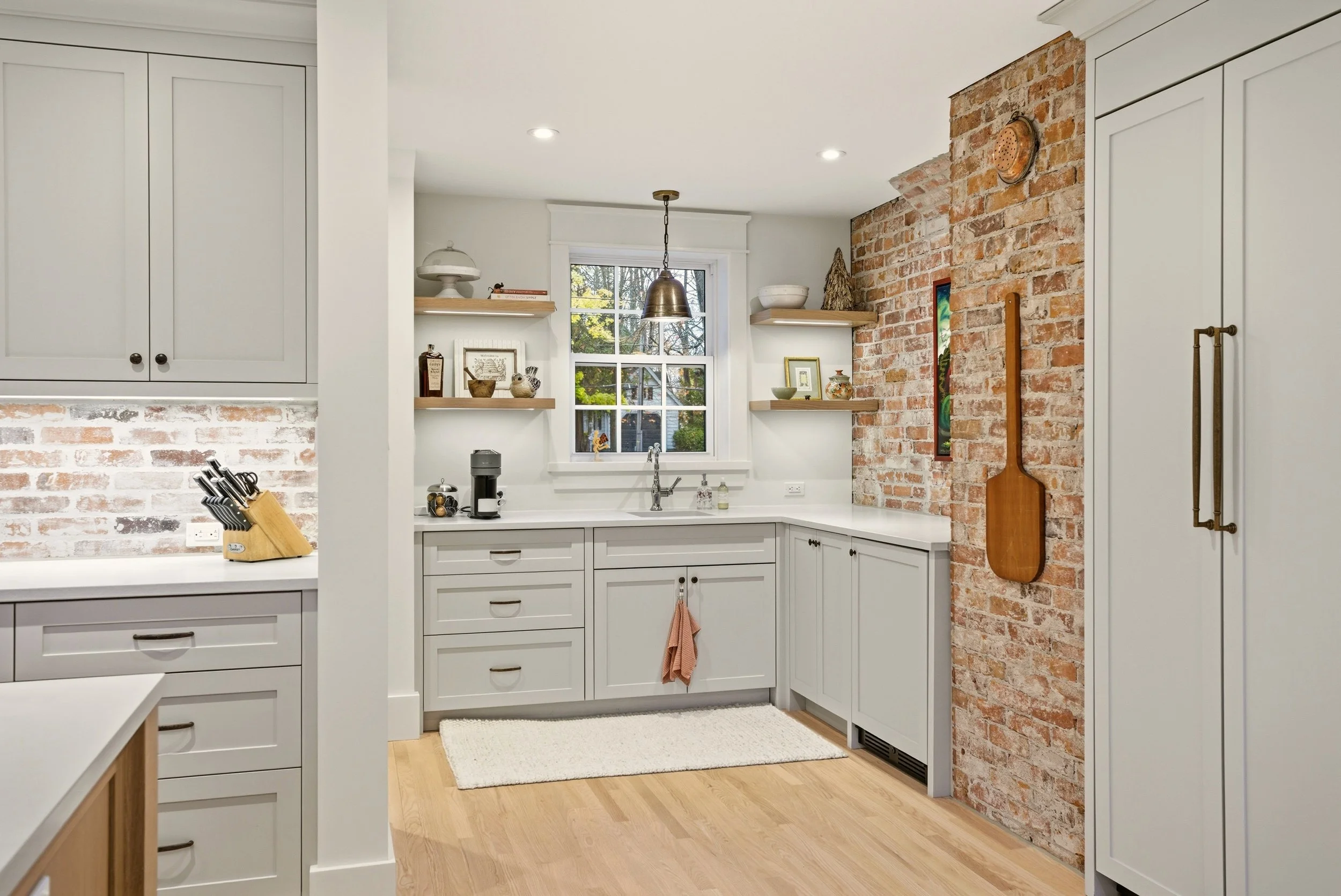 Kitchen with white cabinetry, brick accent wall, wooden paddle hanging on the brick wall, open shelves with decor, window above sink, light wood flooring, small white rug.