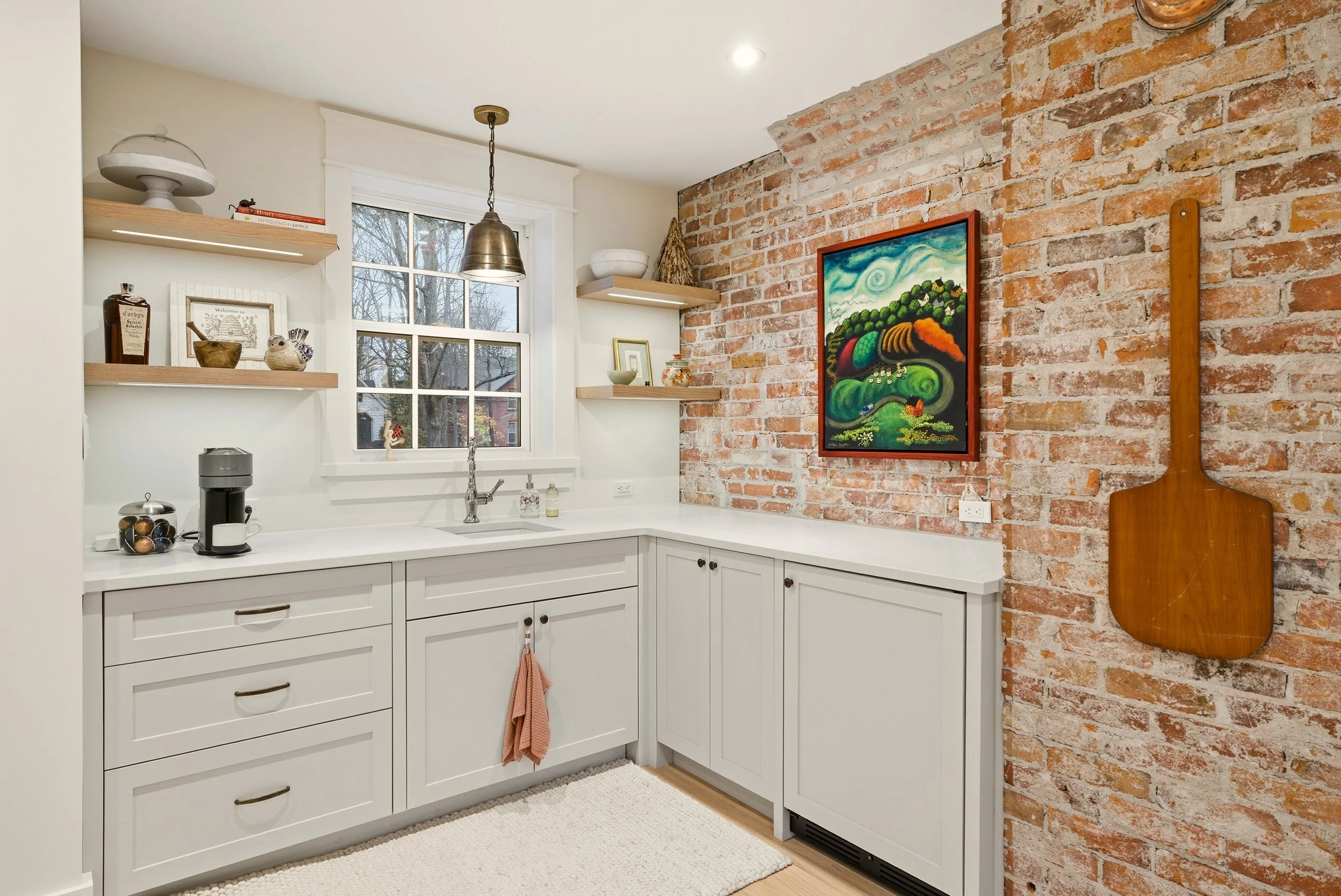 Kitchen with white cabinets, a brick wall, and a window. Decor includes shelves with various items, a framed painting, and a wooden paddle hanging on the brick wall.