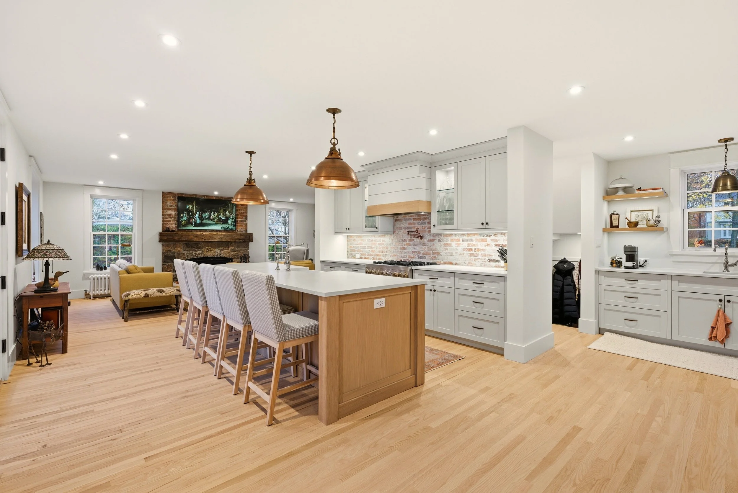 Open-concept kitchen and living room in Muskoka, built by Conklin's Construction featuring white cabinets, brick backsplash, island with seating, hardwood floors, brick fireplace, and television, with large windows and minimalist decor.