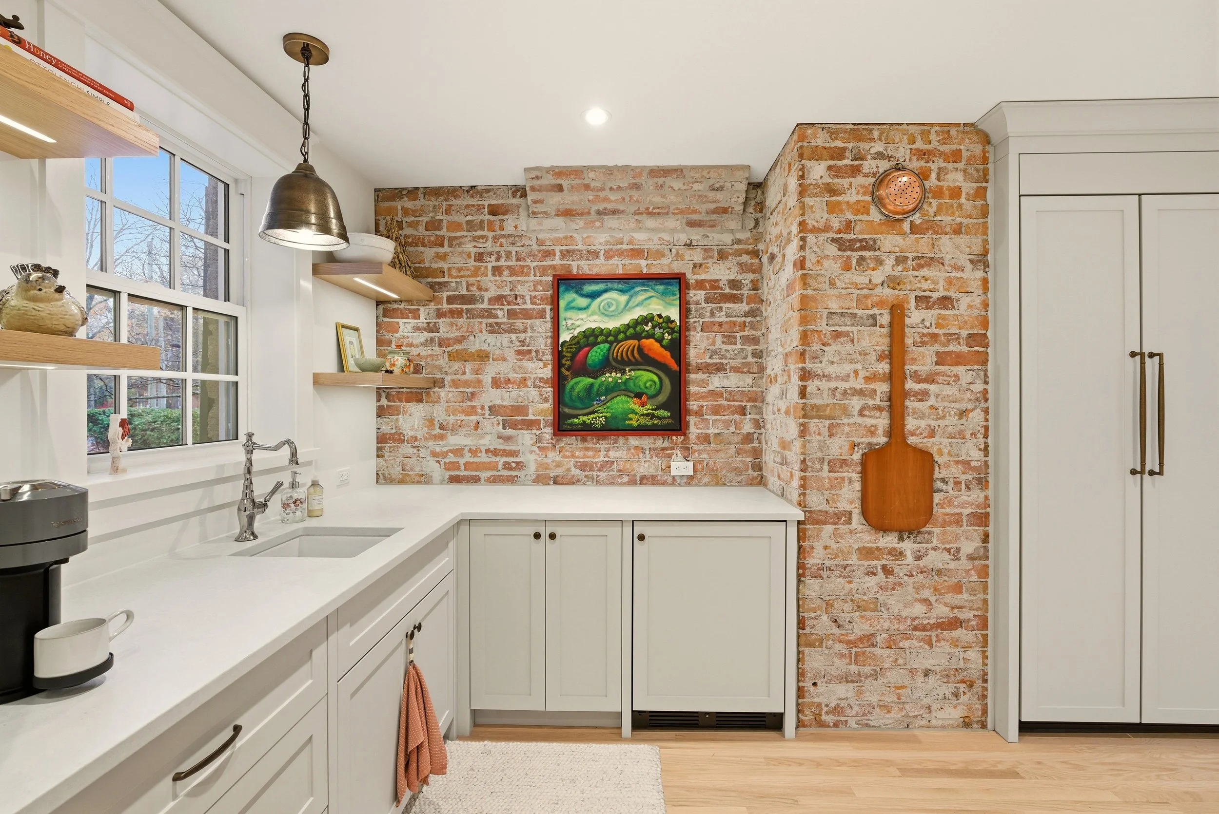 Kitchen with white cabinets, a brick wall, a window with a view outside, shelves with decorative items, a painting on the brick wall, a hanging light fixture, a wooden paddle hanging on the brick wall, and a coffee machine on the counter.