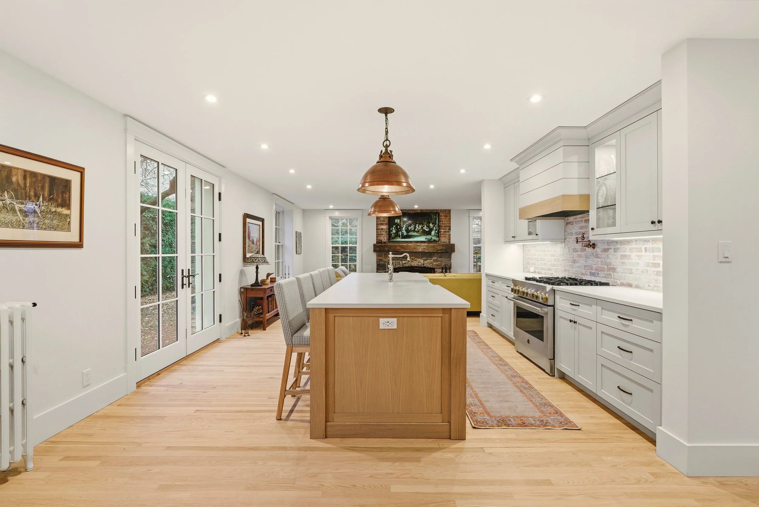 Open-concept kitchen and living area with white cabinets, a kitchen island, hardwood floors, and a brick fireplace in the background. There are large windows and glass doors leading outside, with a few pieces of artwork on the walls.