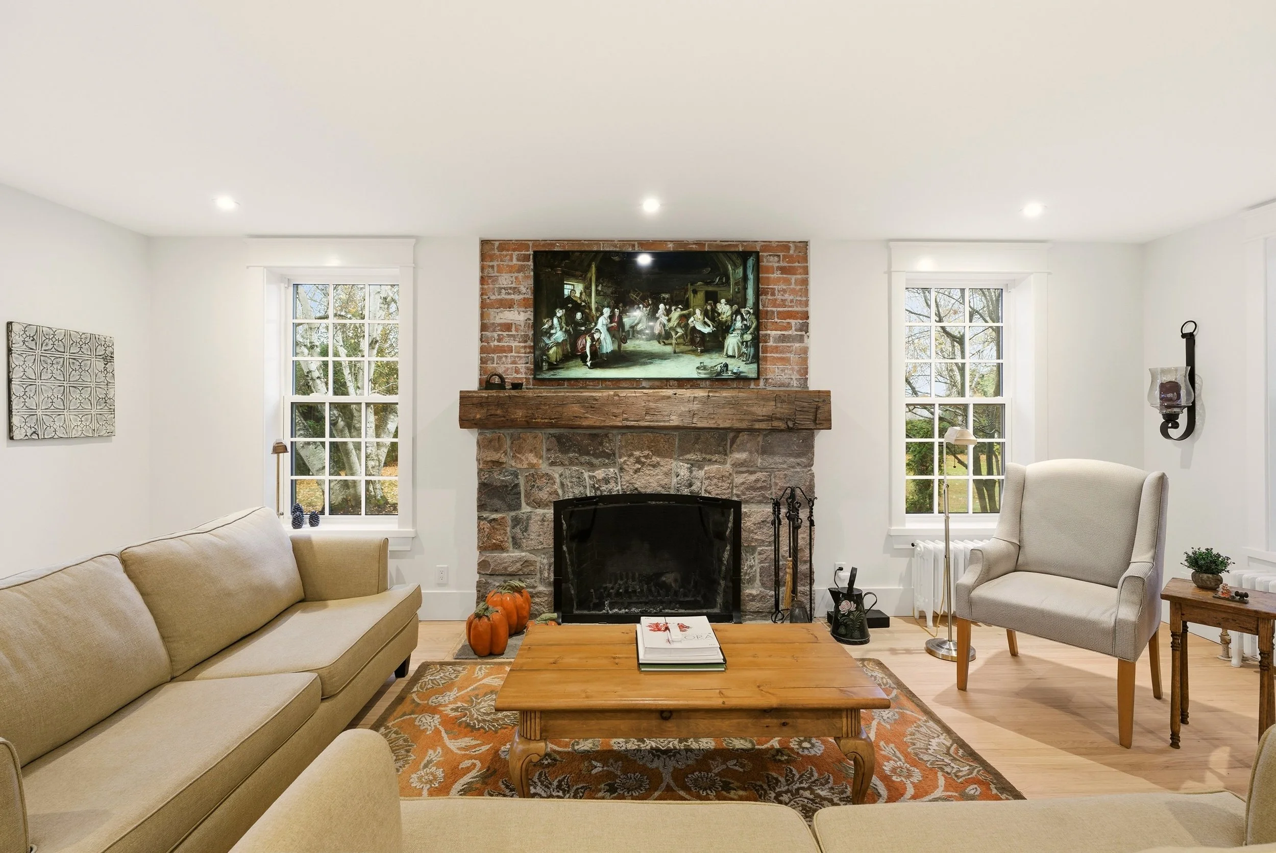Living room with beige sofa, armchair, wooden coffee table, fireplace with stone and brick, large TV above fireplace, windows showing outdoor trees, autumn decor, and neutral walls.