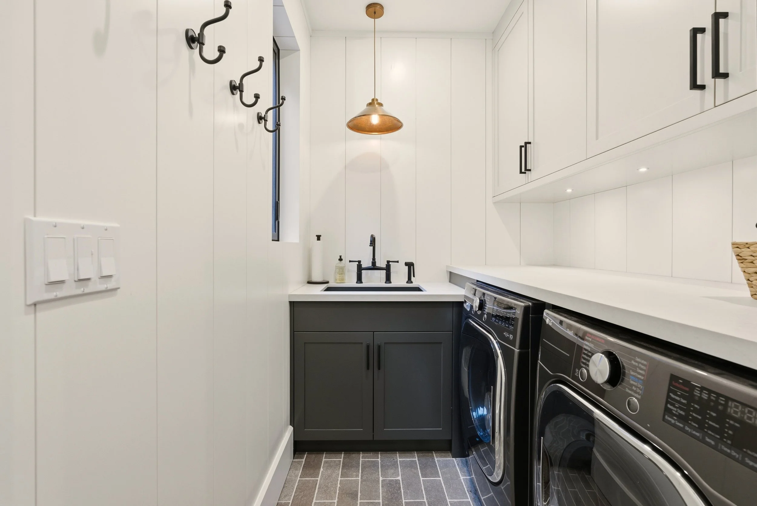 Modern laundry room with white walls, black cabinetry, and a white countertop. There is a black washer and dryer, a small black sink with black fixtures, a pendant light hanging from the ceiling, a narrow window, and wall hooks for hanging items.