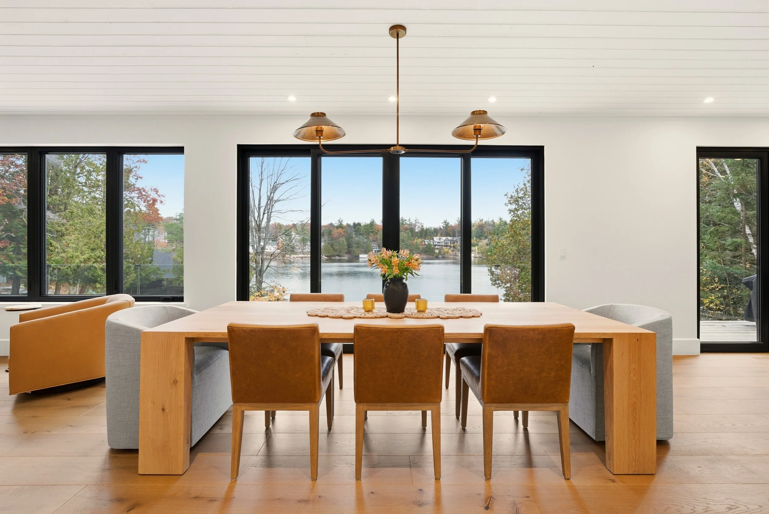 Modern dining room with large windows overlooking a lake, featuring a wooden table, six brown chairs, a vase with flowers, and two yellow candles.