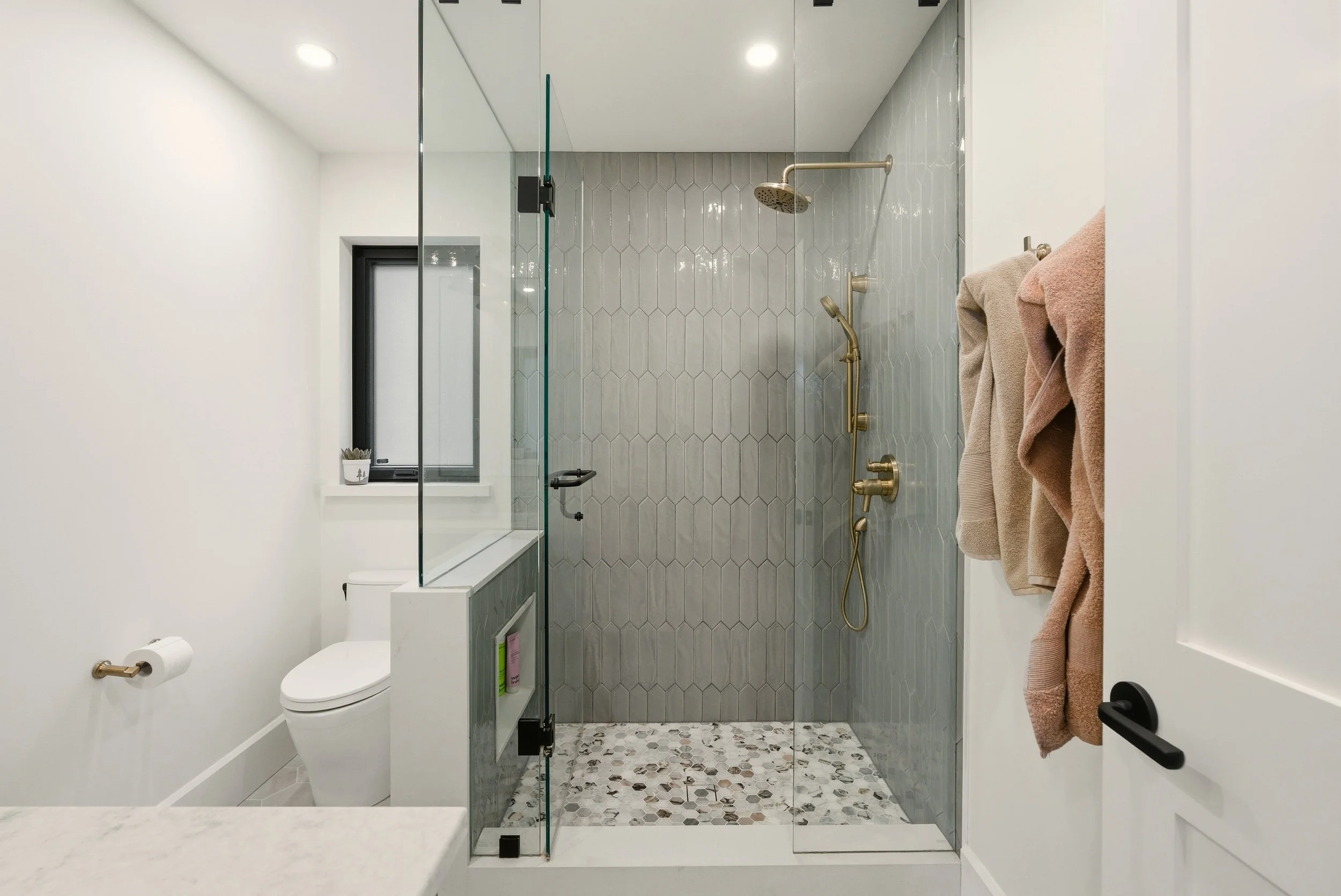Modern bathroom with a glass-enclosed shower area featuring brass fixtures, a toilet, towels hanging on the door, a small window, and a pebble-tile floor in the shower.